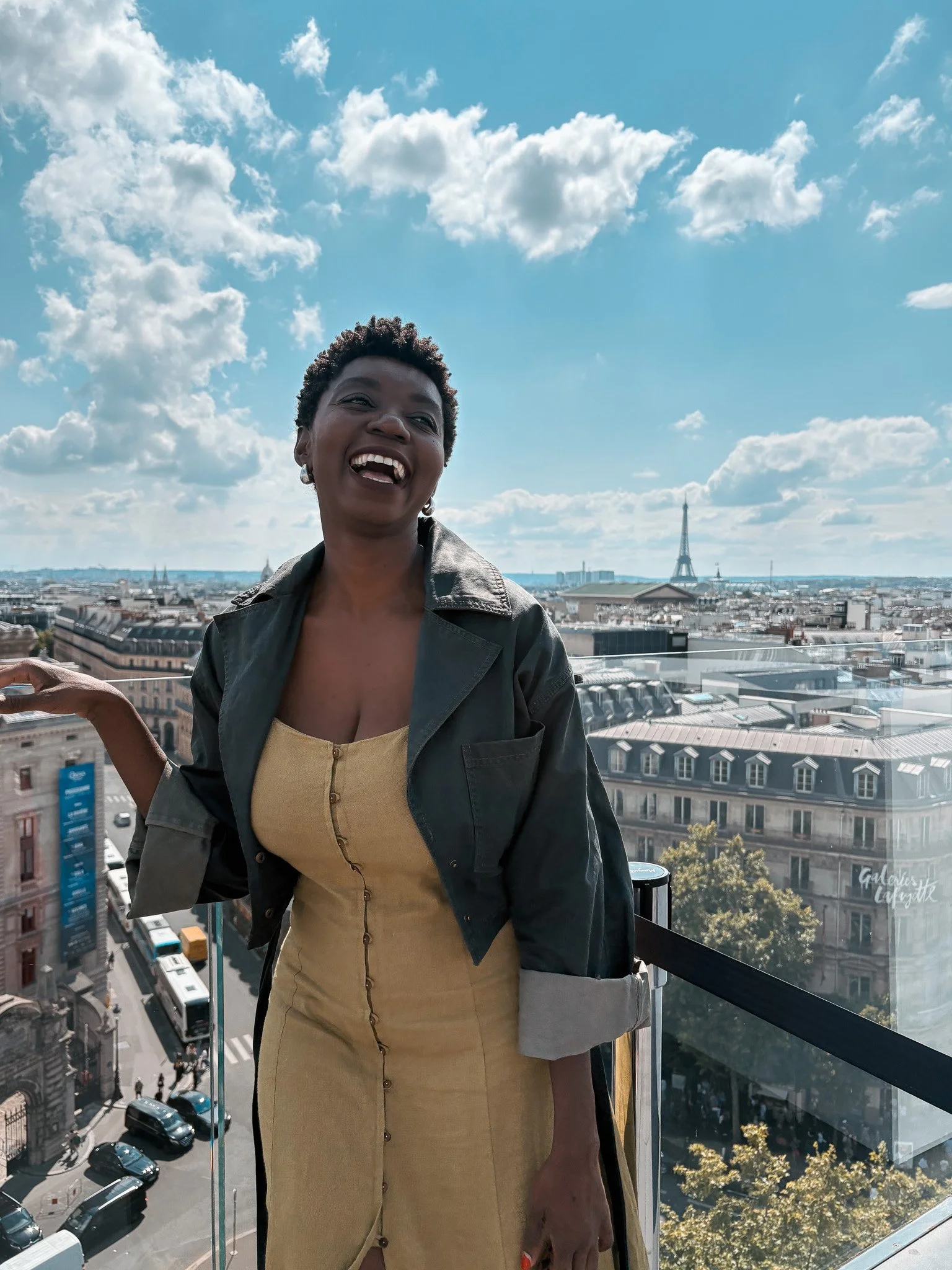 A woman is standing on a balcony in Paris with the Eiffel Tower in the background. She is smiling and wearing a yellow dress with a green jacket. The sky is blue with scattered clouds.