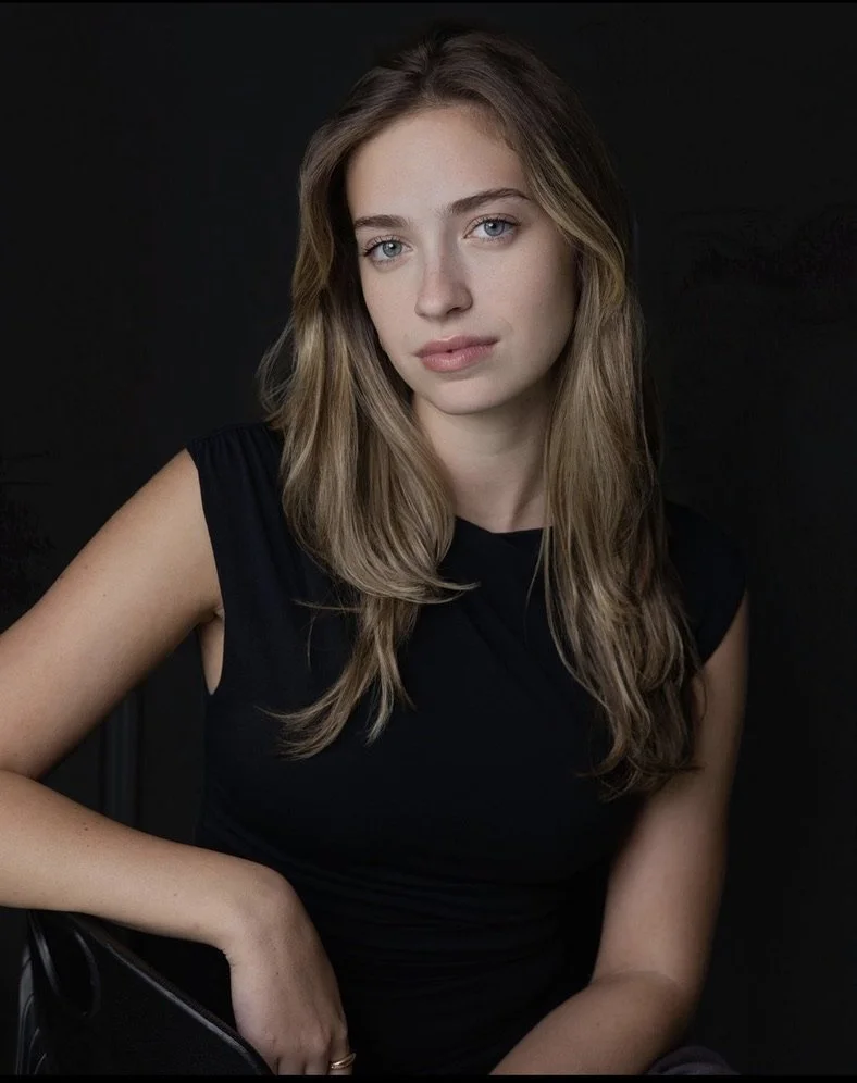 A young woman with long wavy blonde hair and blue eyes, wearing a black sleeveless top, posing against a dark background.