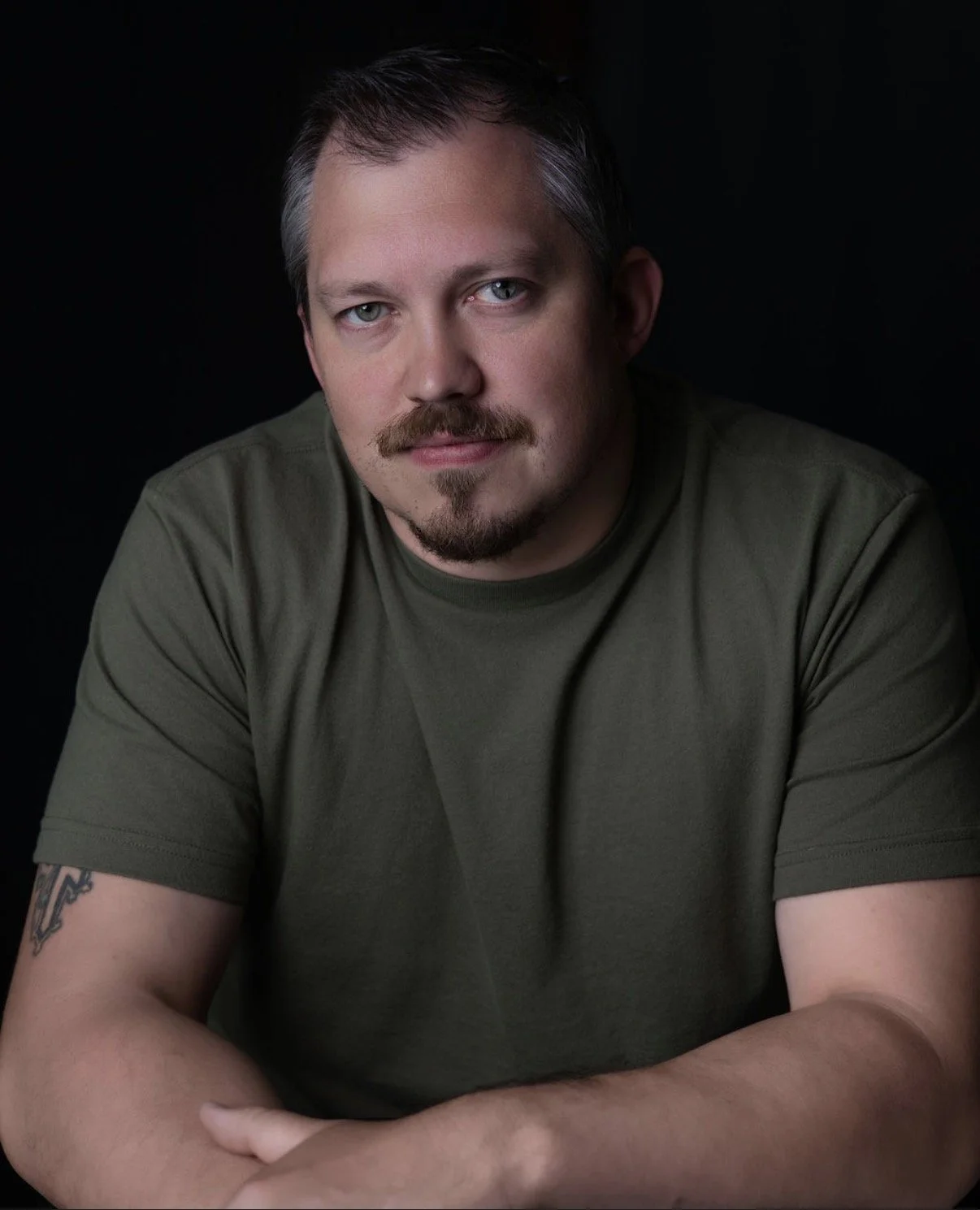 A man with short dark hair, a mustache, and a goatee, wearing a green t-shirt with tattoos visible on his right arm, sitting against a dark background.