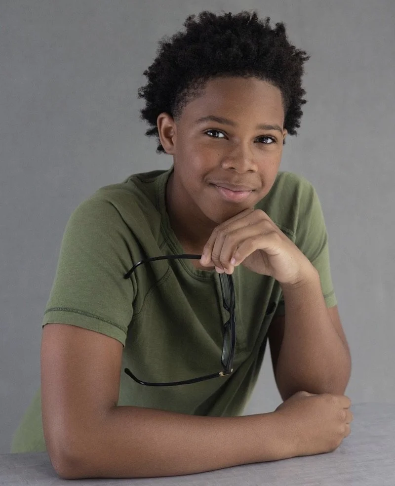A young boy with curly black hair, wearing an olive green t-shirt, sitting at a table with sunglasses hanging from his shirt collar, smiling at the camera.