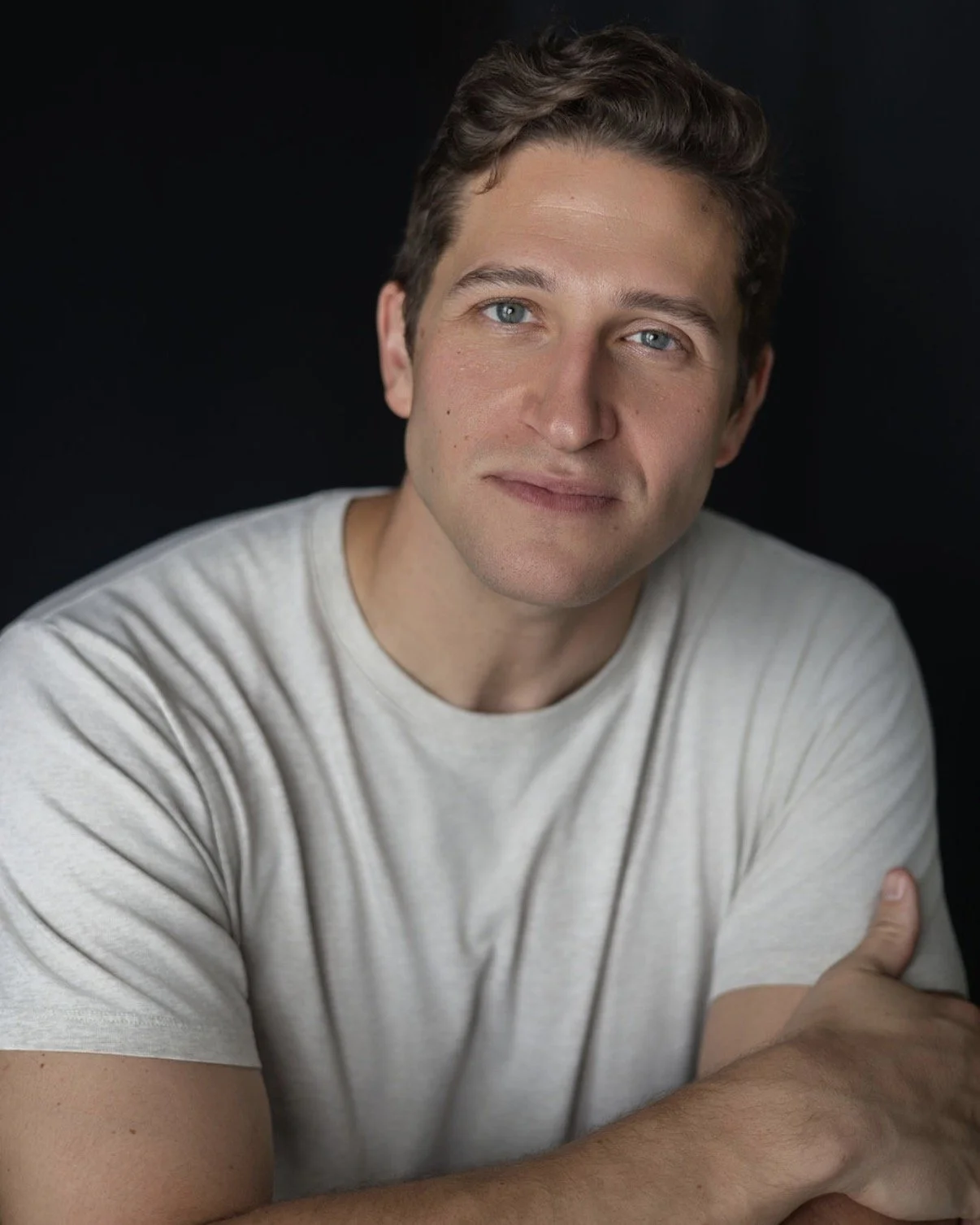 Headshot of a man with blue eyes and brown hair, wearing a light-colored t-shirt, against a dark background.