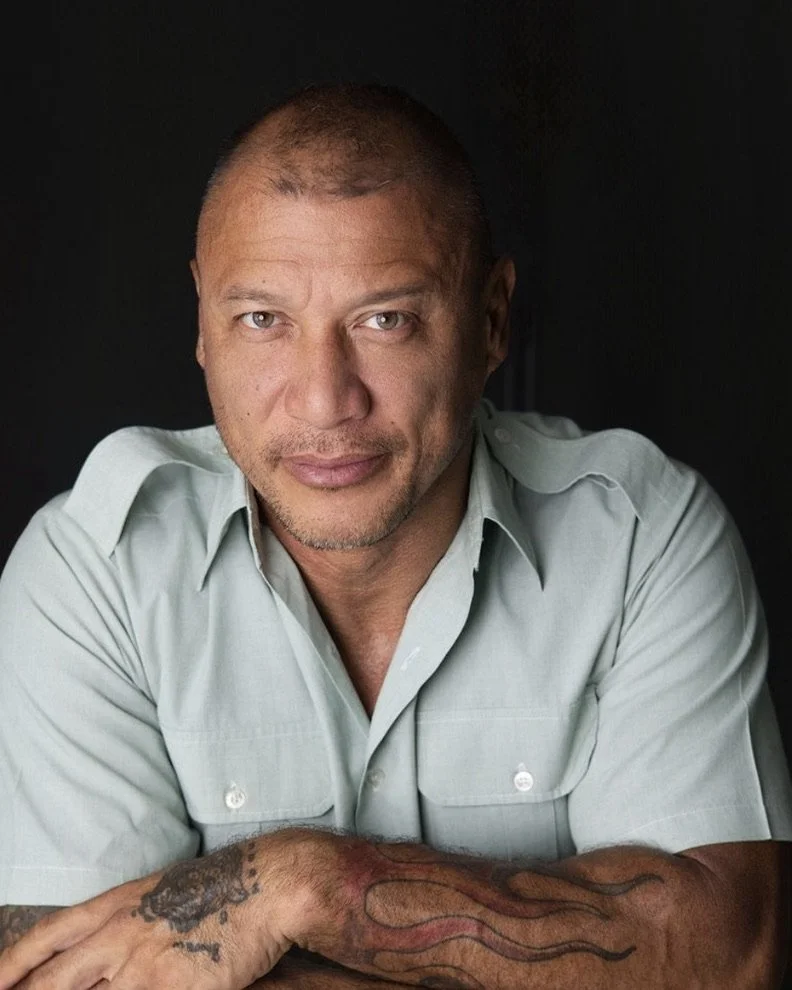 A middle-aged man with short hair and a light beard, wearing a light green button-up shirt, leaning forward with crossed arms, looking directly at the camera against a dark background.
