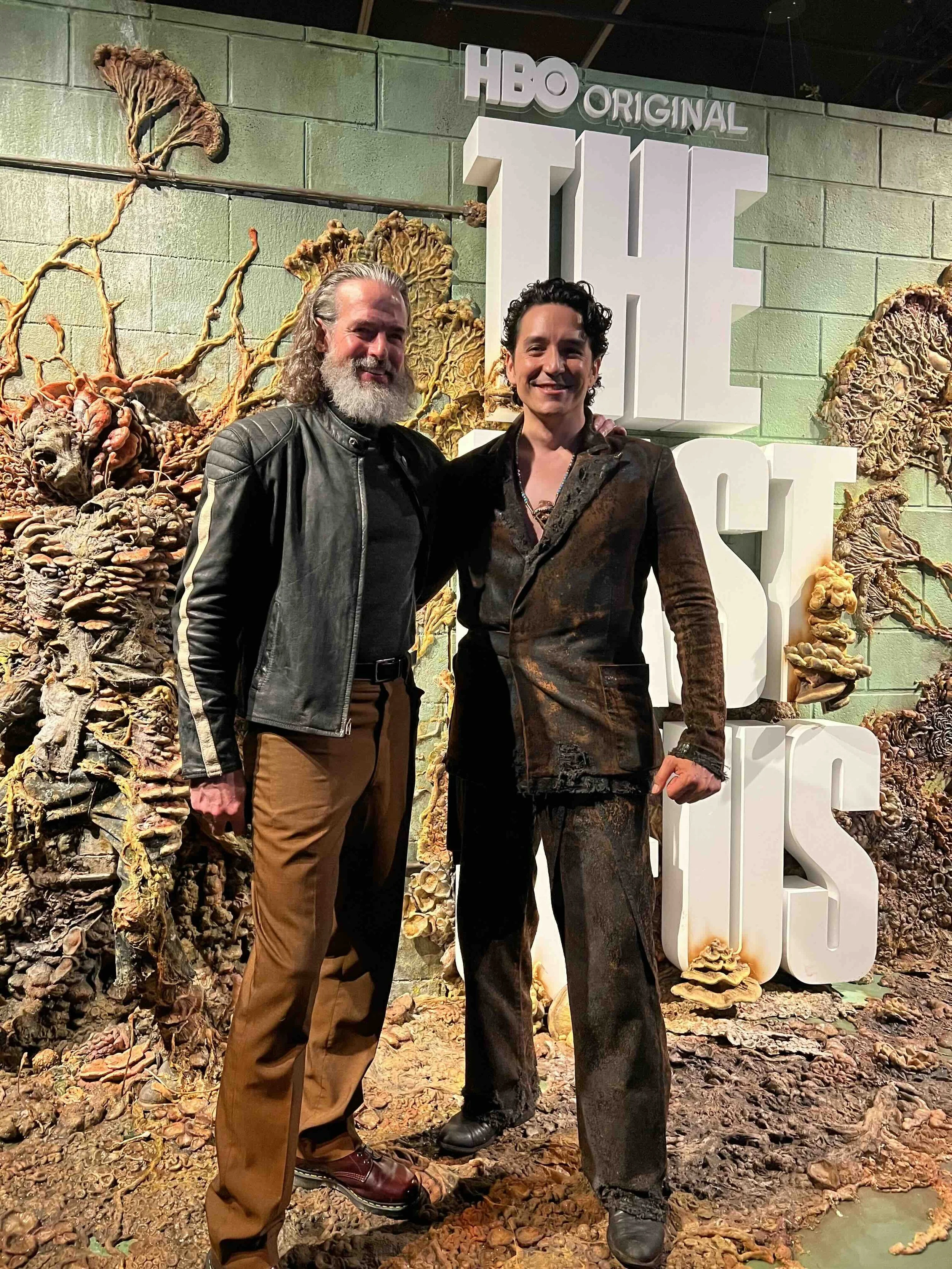 Two men standing in front of a textured green wall with large white letters spelling 'THE OFFICE' and a sign that says 'HBO ORIGINAL.' One man has long gray hair and a beard, wearing a black leather jacket and brown pants. The other man has short dark curly hair and is wearing a patterned suit. They are smiling, with their arms around each other.