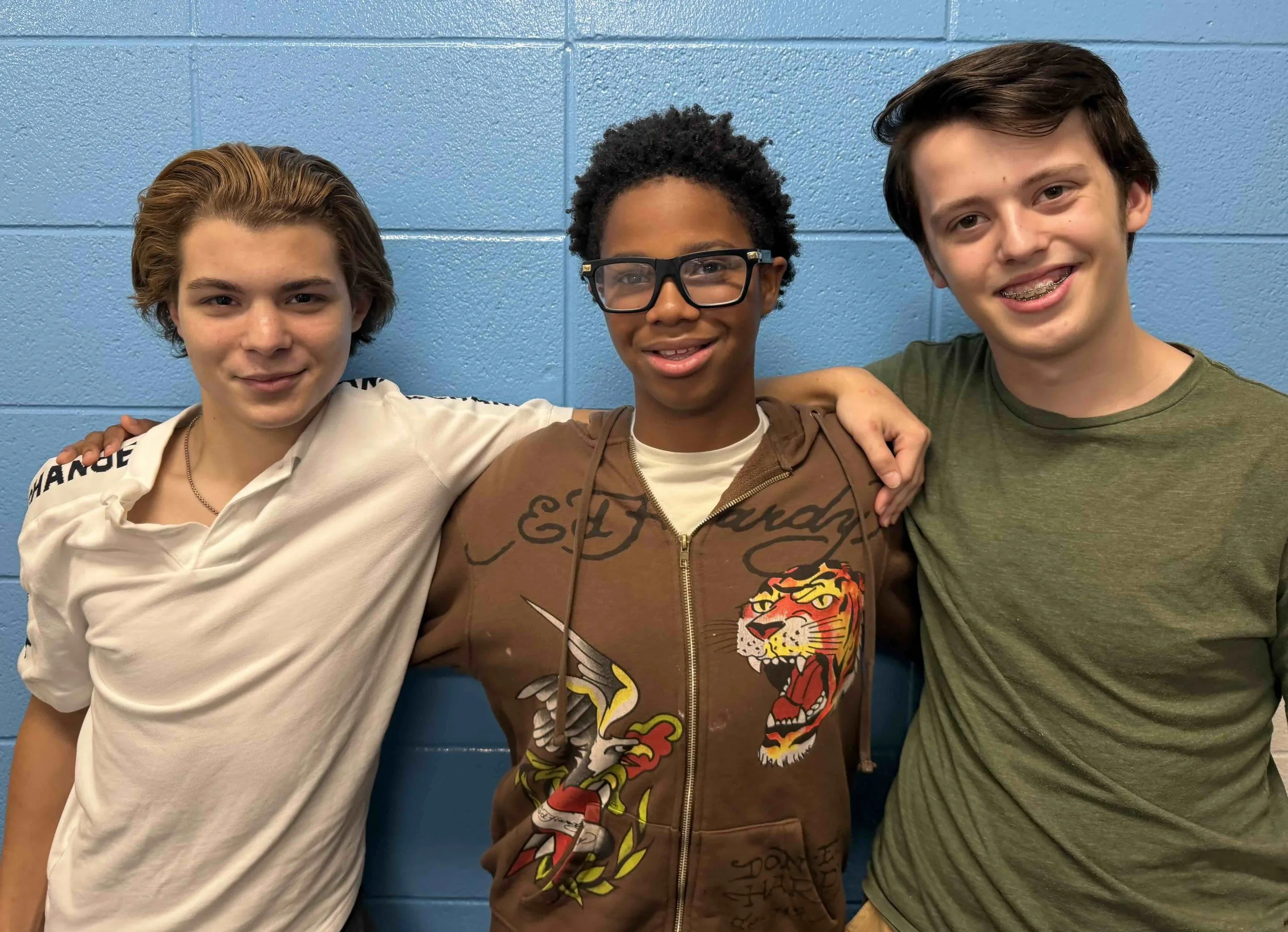 Three boys standing side by side in front of a blue wall, with their arms around each other, smiling at the camera.