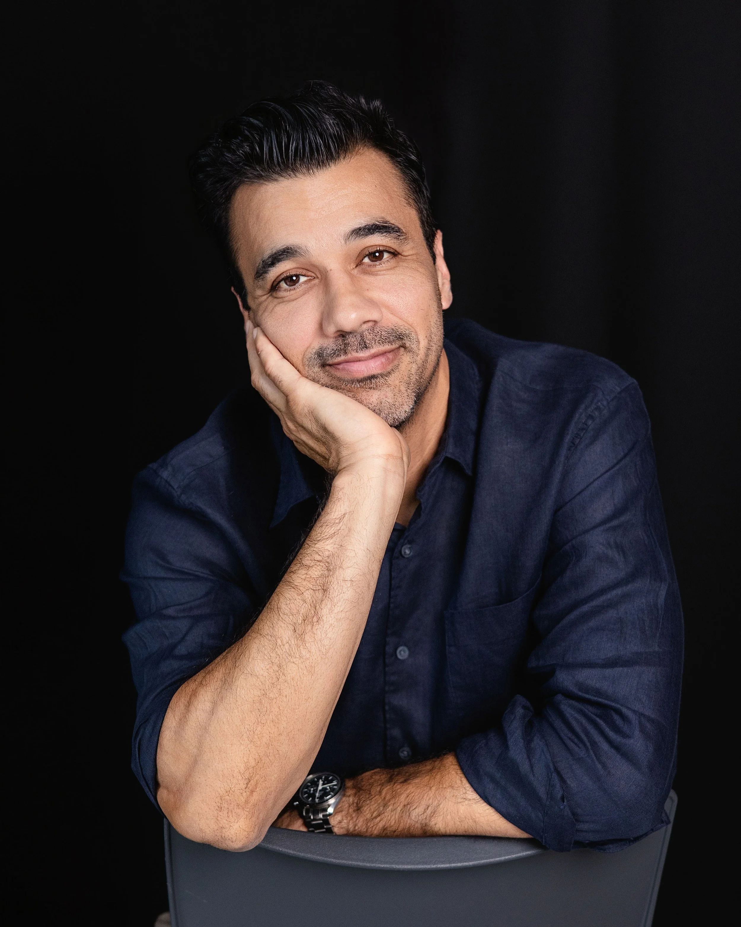 A man with dark hair and a beard, wearing a dark blue shirt and a watch, resting his chin on his hand while smiling at the camera against a black background.
