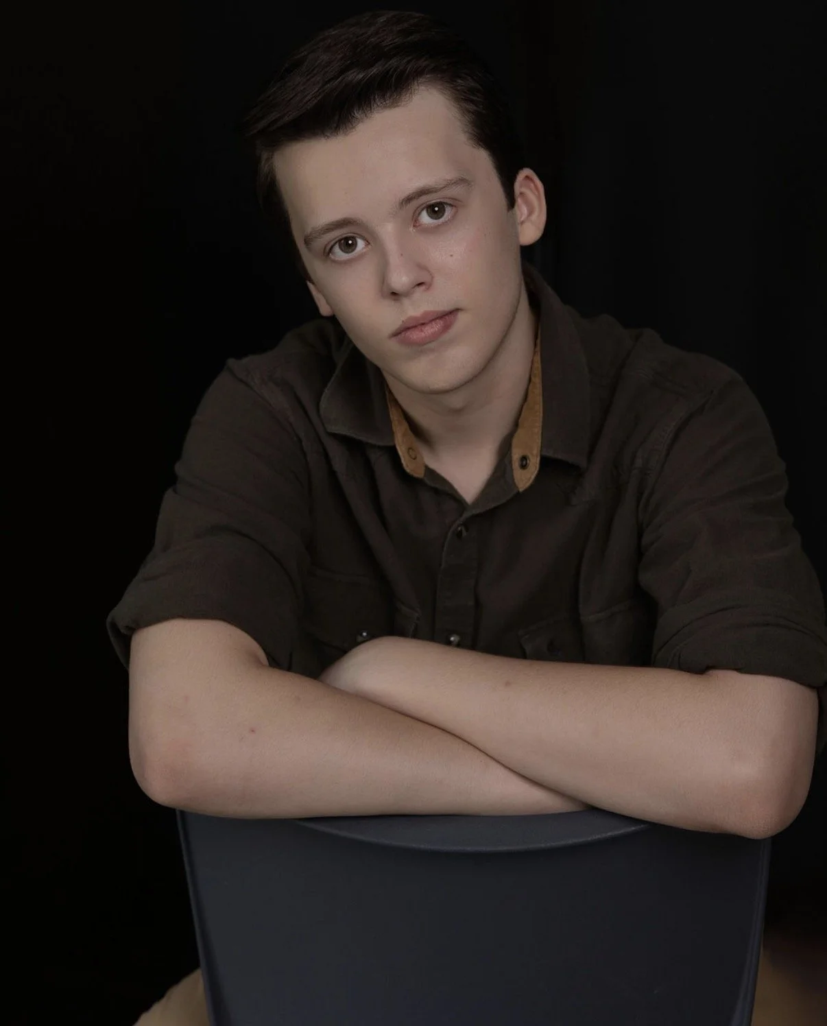 A young man with dark hair and light skin sits with his arms crossed on a dark chair, looking at the camera with a neutral expression, against a black background.