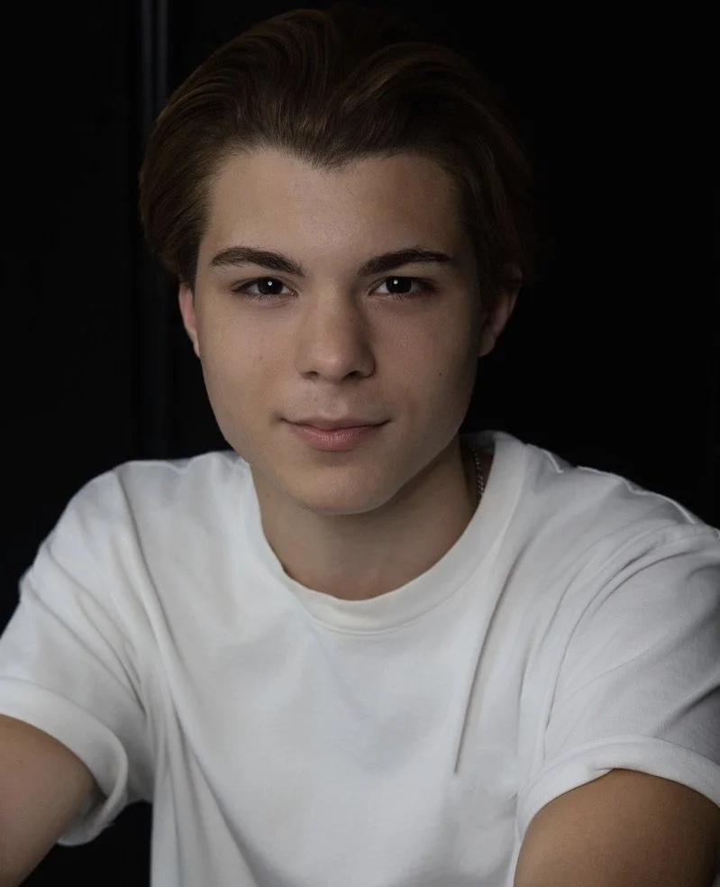 Portrait of a young man with light brown hair wearing a white t-shirt against a black background.