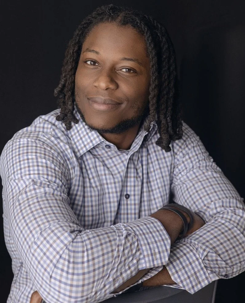 Portrait of a man with dreadlocks, wearing a checkered shirt and a wristwatch, smiling against a dark background.