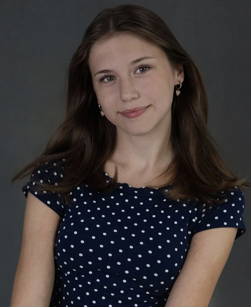 A young woman with long brown hair and light skin, wearing a navy blue polka dot dress, smiling slightly, with heart-shaped earrings, set against a dark gray background.