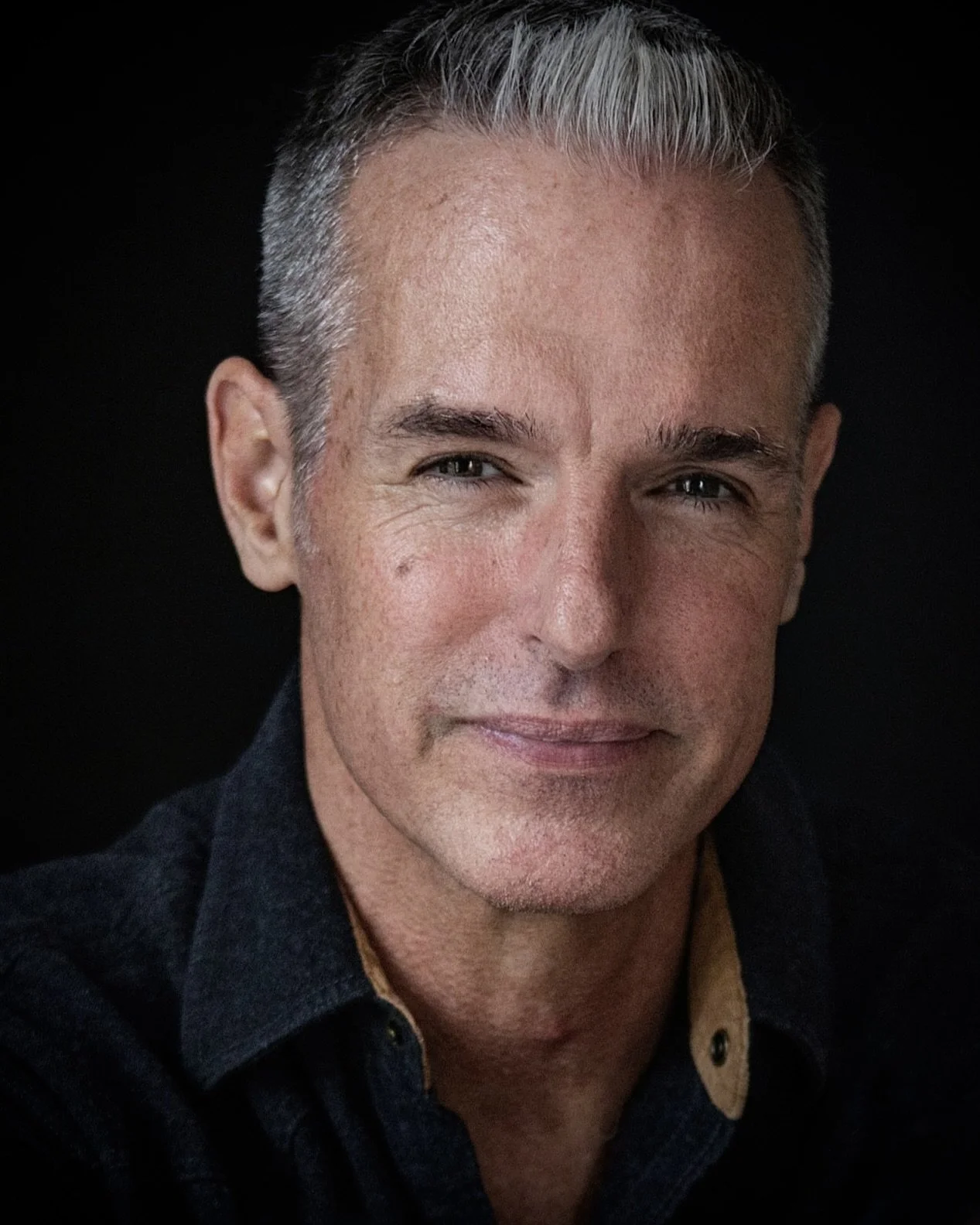 Close-up of a middle-aged man with short gray hair, blue eyes, and a slight smile, wearing a dark shirt with a tan collar against a black background.