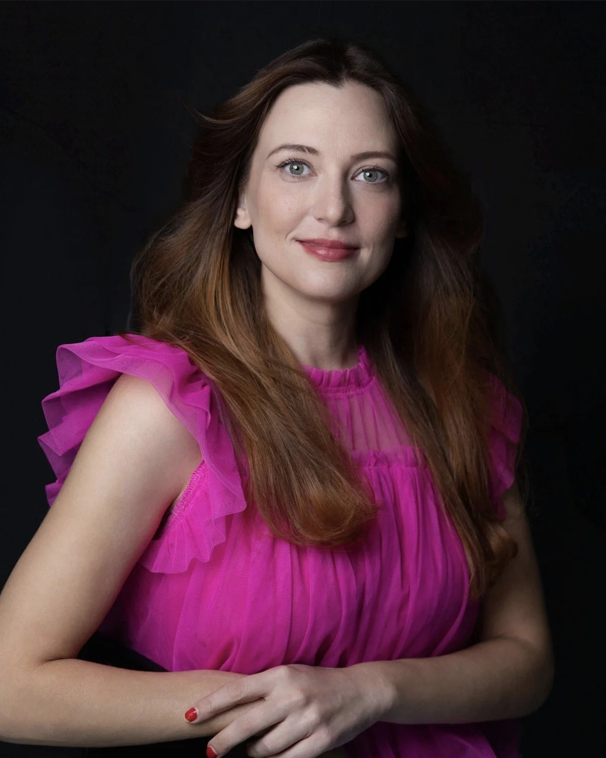 Portrait of a woman with long, wavy brown hair, wearing a bright pink, ruffled sleeveless top, against a dark background.