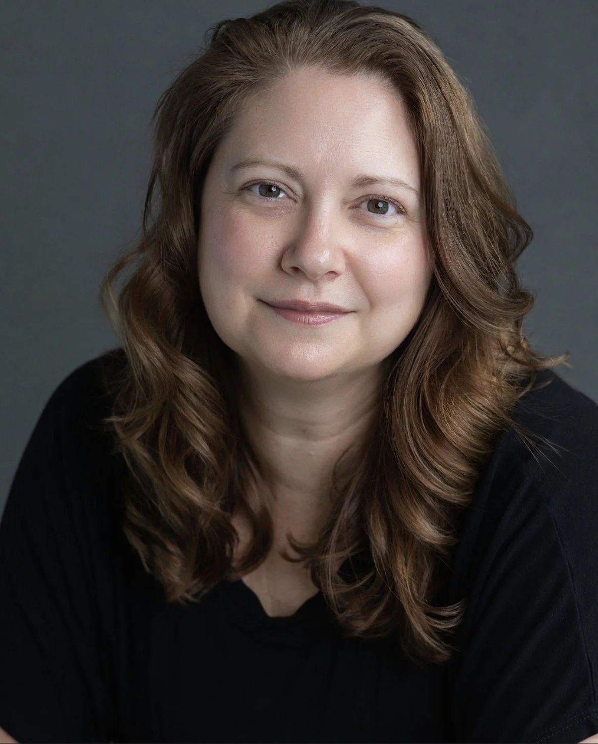 A woman with shoulder-length wavy brown hair, light skin, and wearing a black top, looking at the camera with a slight smile against a plain dark gray background.