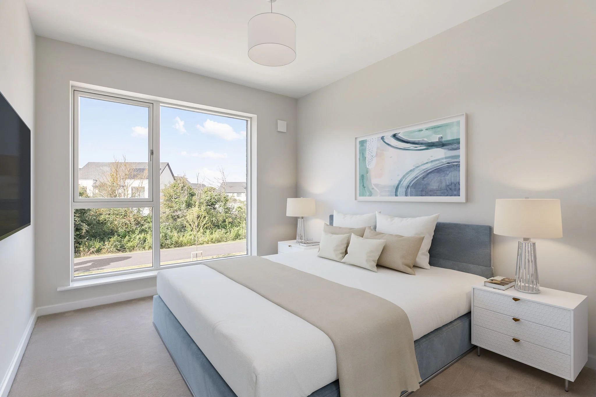 A neatly made bed with white and beige pillows and a beige throw blanket in a modern, minimalist bedroom. Two white nightstands with matching lamps are on either side of the bed. A large abstract painting hangs above the bed. A window shows a sunny o