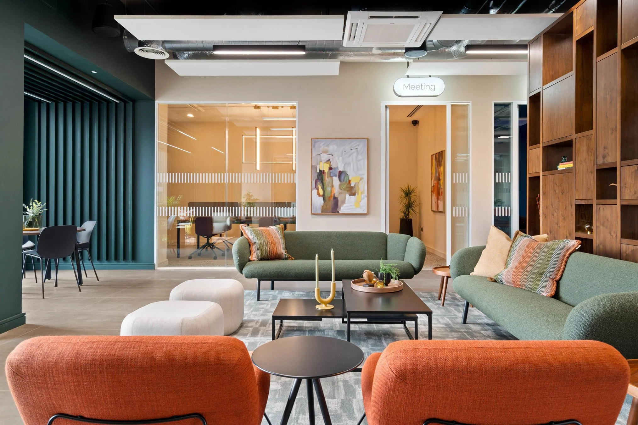 Modern office lounge area with green and orange sofas, wooden bookshelves, abstract artwork, and a meeting room with glass walls in the background.