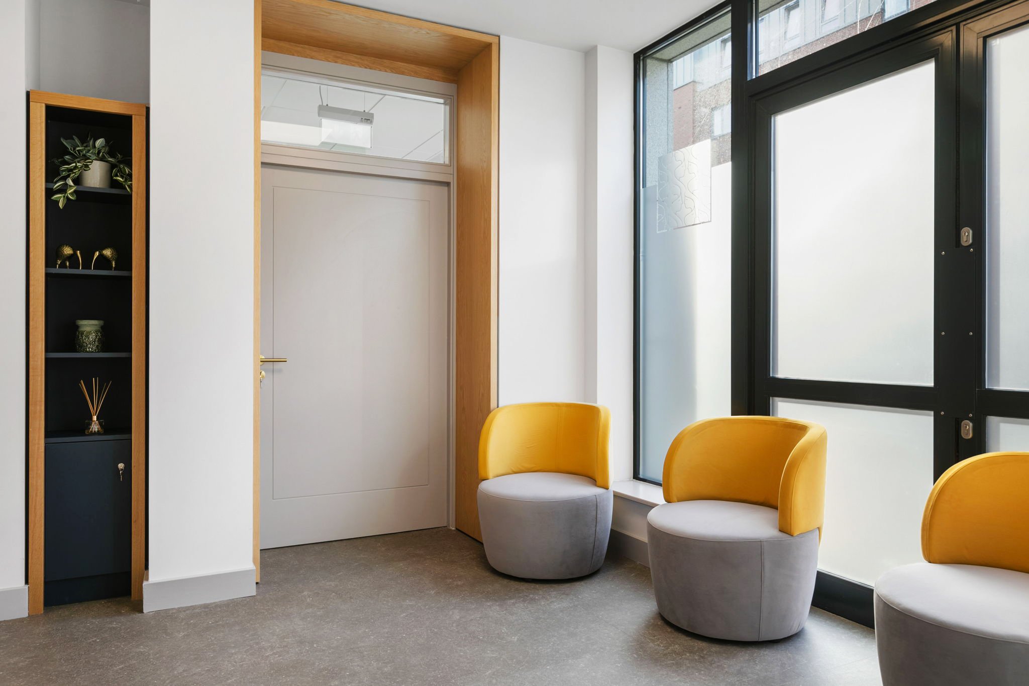 Modern waiting area with two yellow and gray chairs near large glass windows and a black shelf with decorative items on the left.