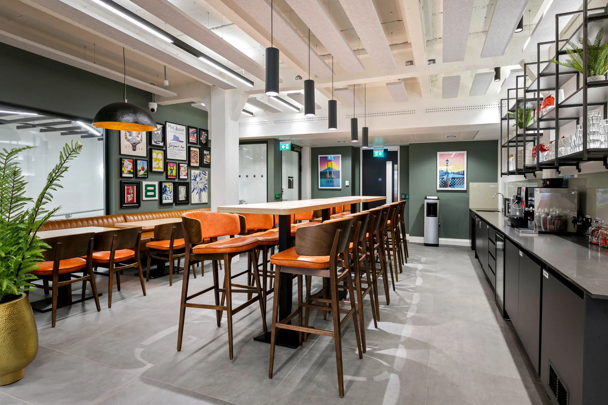 Modern cafe or lounge area with bar-height chairs, a high table, artwork on the wall, and a coffee station with glasses and supplies.