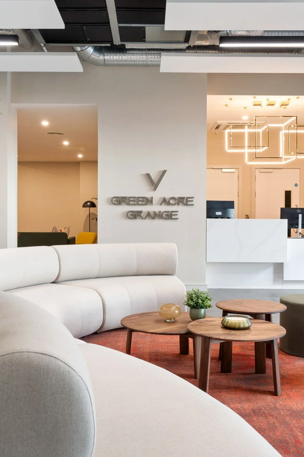 Modern hotel lobby with white sofas, wooden tables, and a sign on the wall that reads 'Green Acre Grange' with a check mark logo. There is a reception desk in the background and contemporary lighting fixtures.