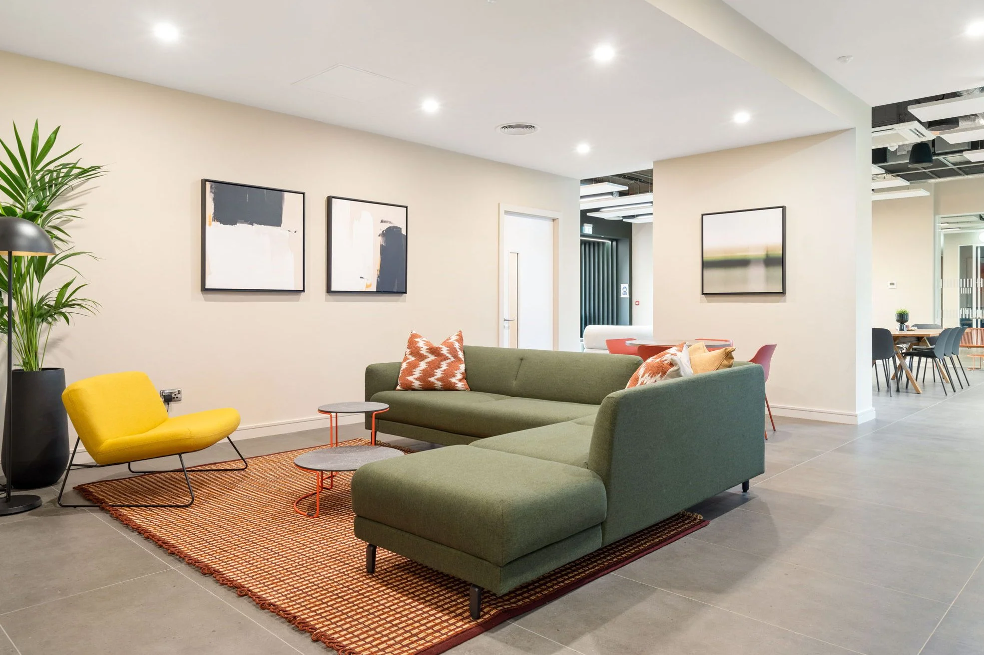 Modern hotel lobby or lounge area with a green sectional sofa, yellow chair, and colorful pillows, artwork on the walls, and a large potted plant.
