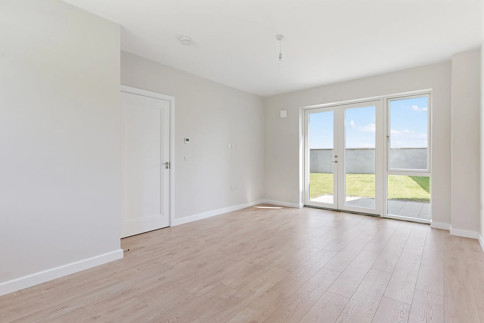 Empty living room with light wood flooring, white walls, a white door, and glass sliding doors leading to a grassy outdoor area.