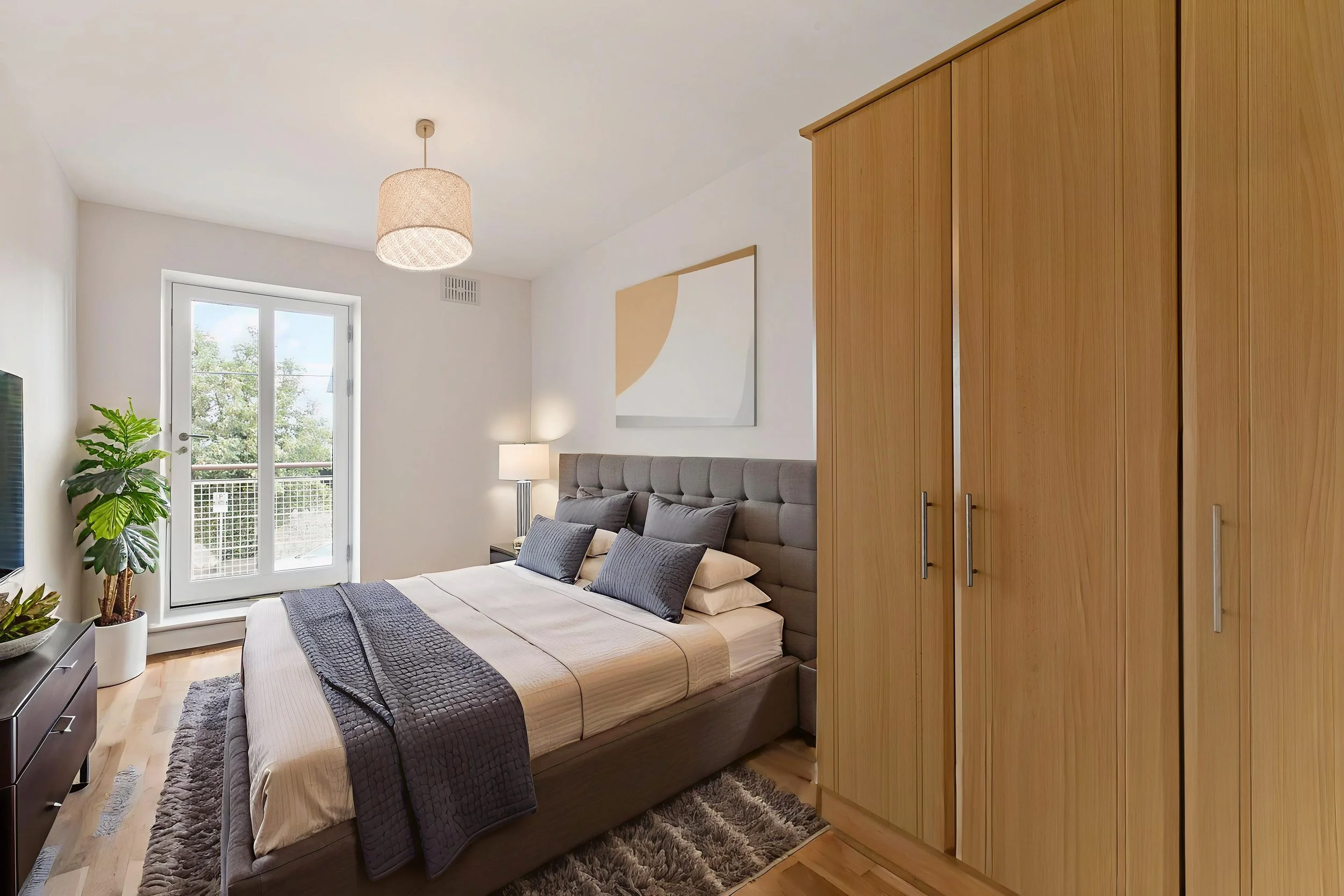 Bedroom with large bed, gray upholstered headboard, multiple pillows, beige and gray bedding, wooden furniture, a large wardrobe, a nightstand with a lamp, a potted plant, a door leading to a balcony, and abstract wall art.