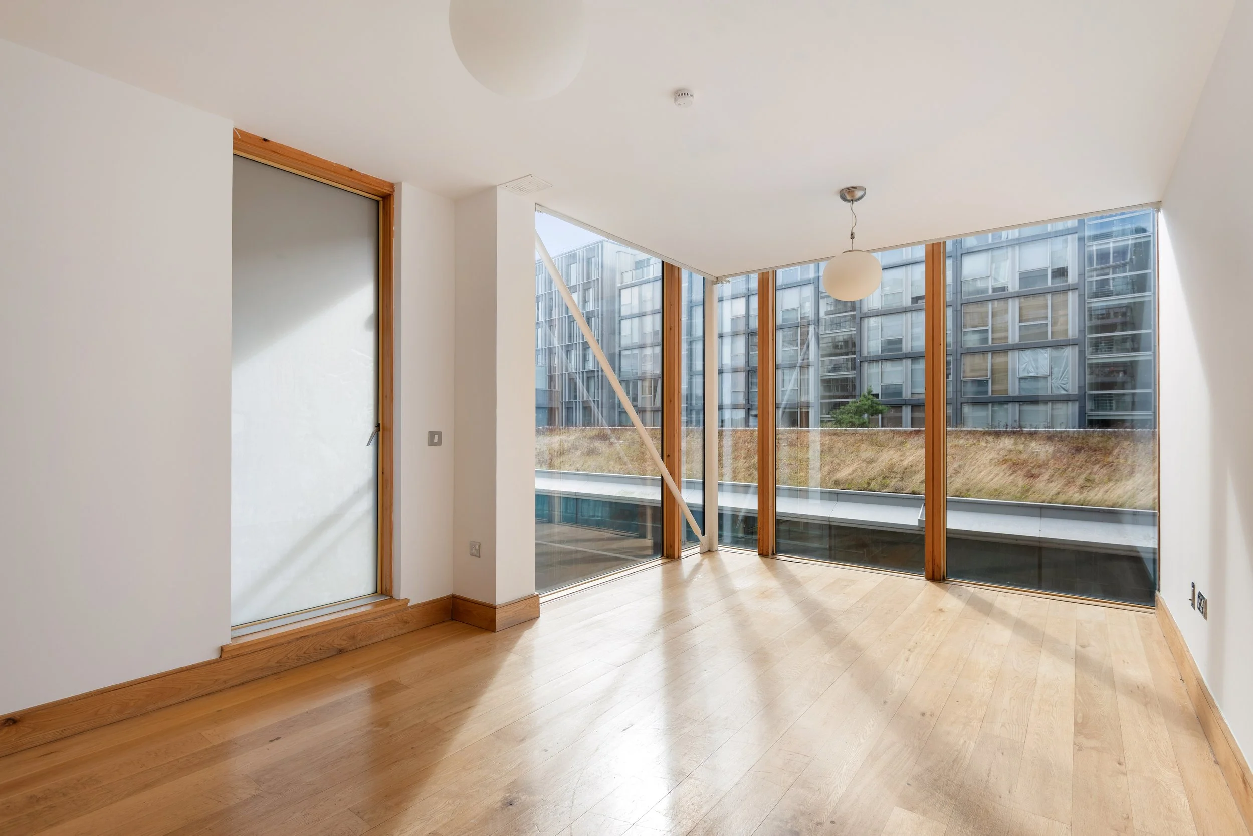 Empty room with large glass windows, wooden floors, and a door with frosted glass, overlooking a grassy hillside and modern apartment buildings.