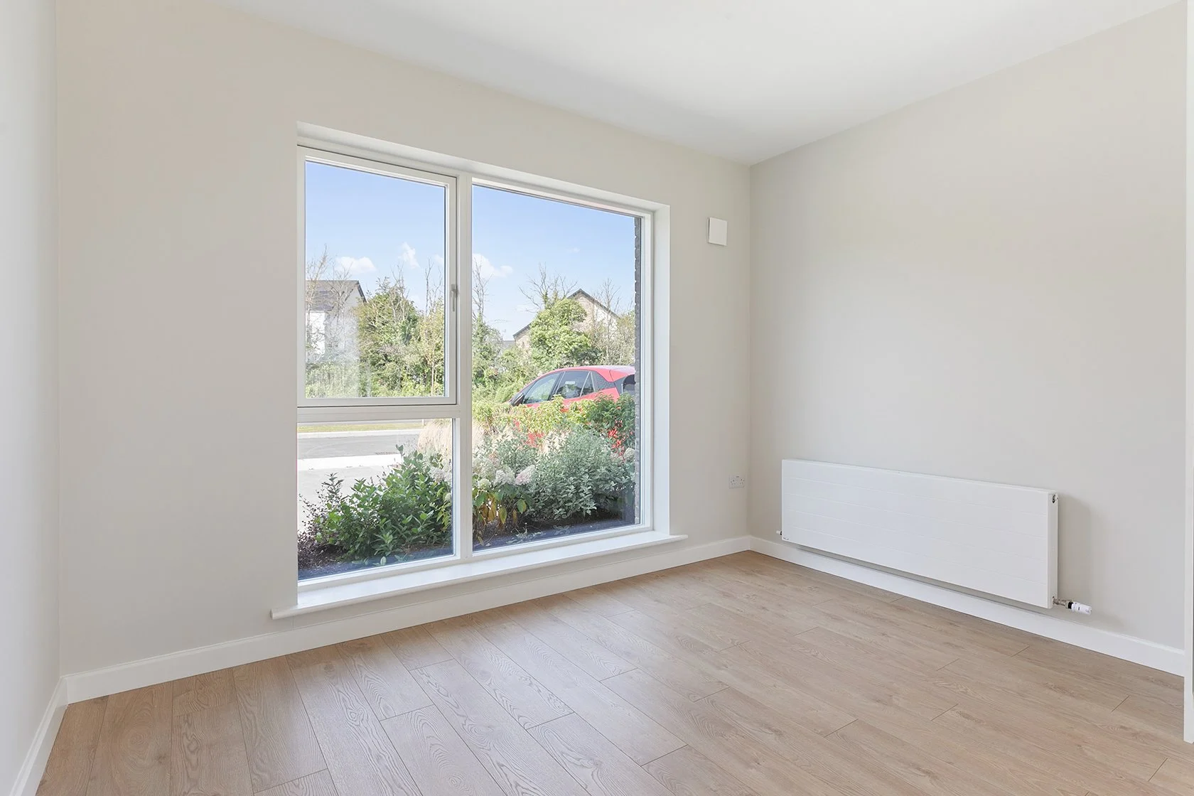 Empty room with large window, white walls, wooden floor, and radiator.
