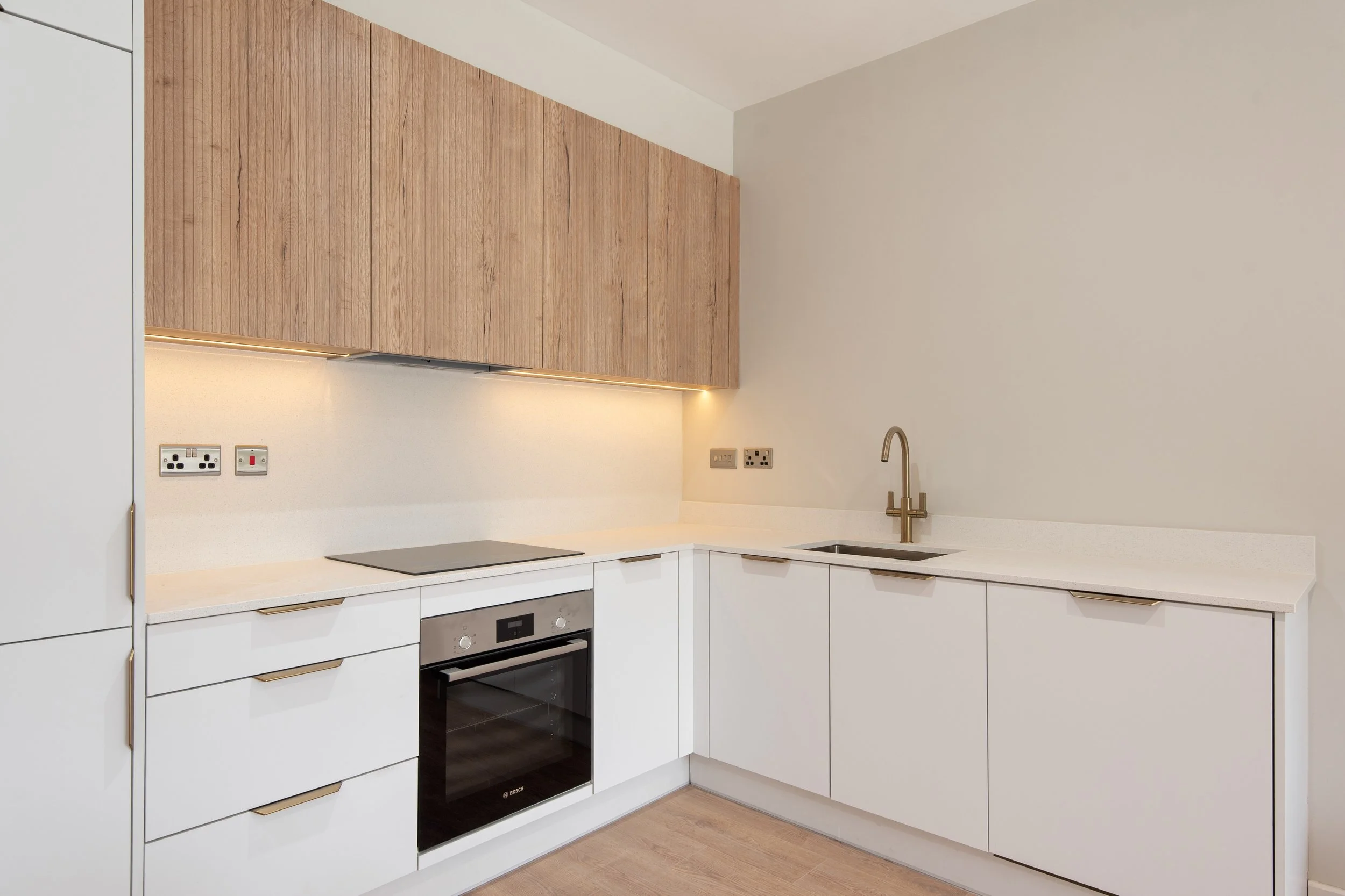 Modern kitchen with white cabinets, a built-in oven, a stovetop, a sink with a brass faucet, and wooden upper cabinets with recessed lighting.