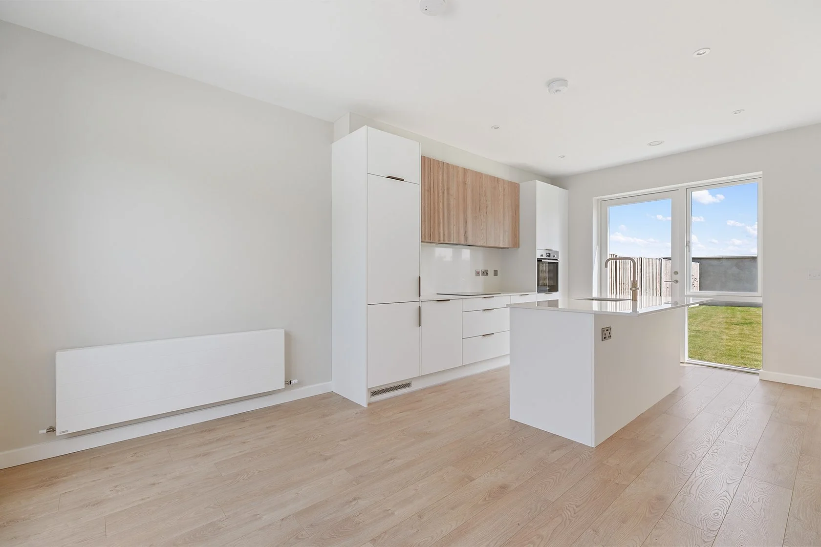 Empty modern kitchen with white cabinets, a wooden cabinet section, built-in oven, and a white kitchen island near large glass doors leading outside, natural light, light wood flooring, ceiling lights.