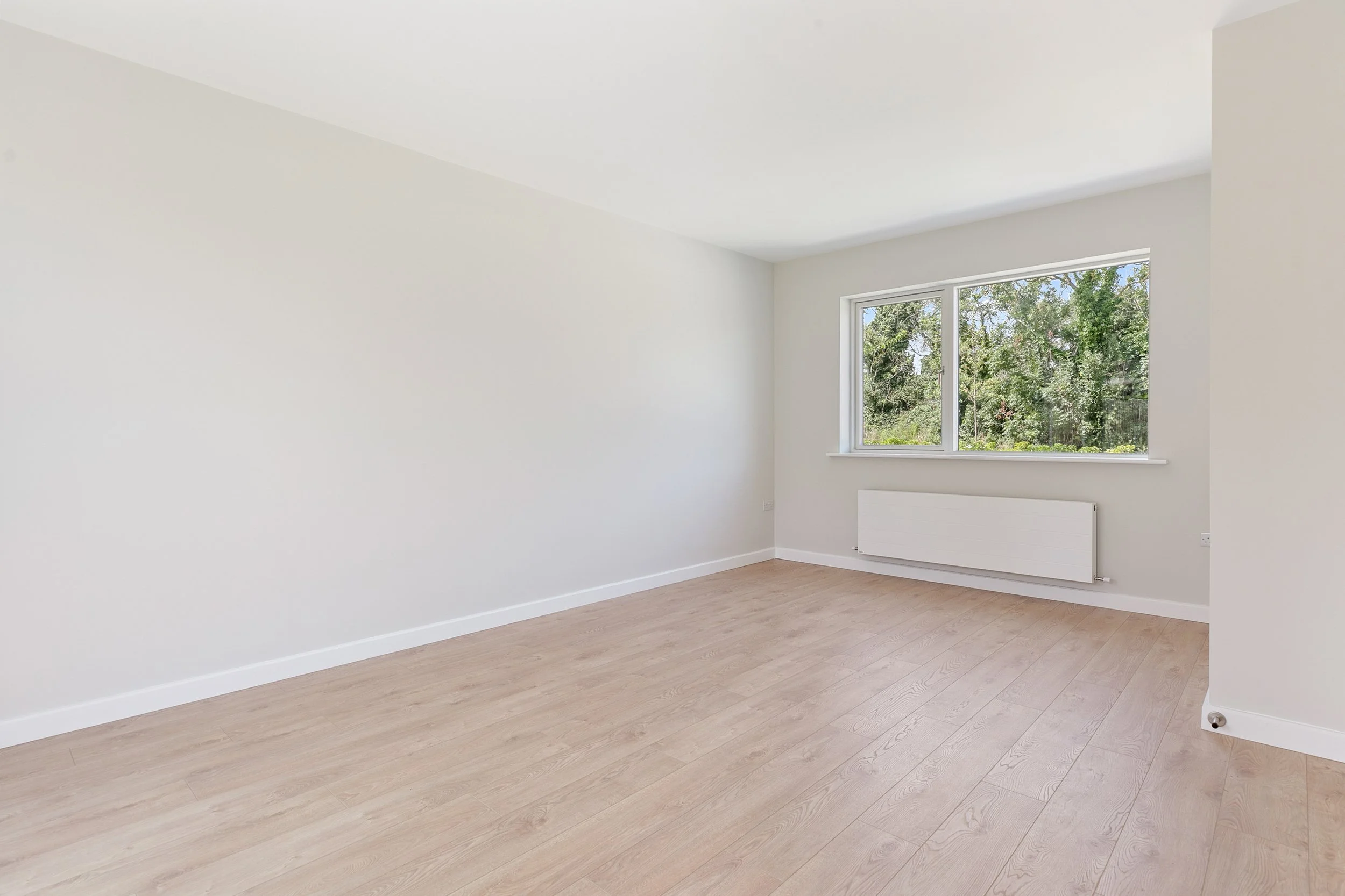 Empty room with white walls, large window, light wood flooring, and radiator beneath window.