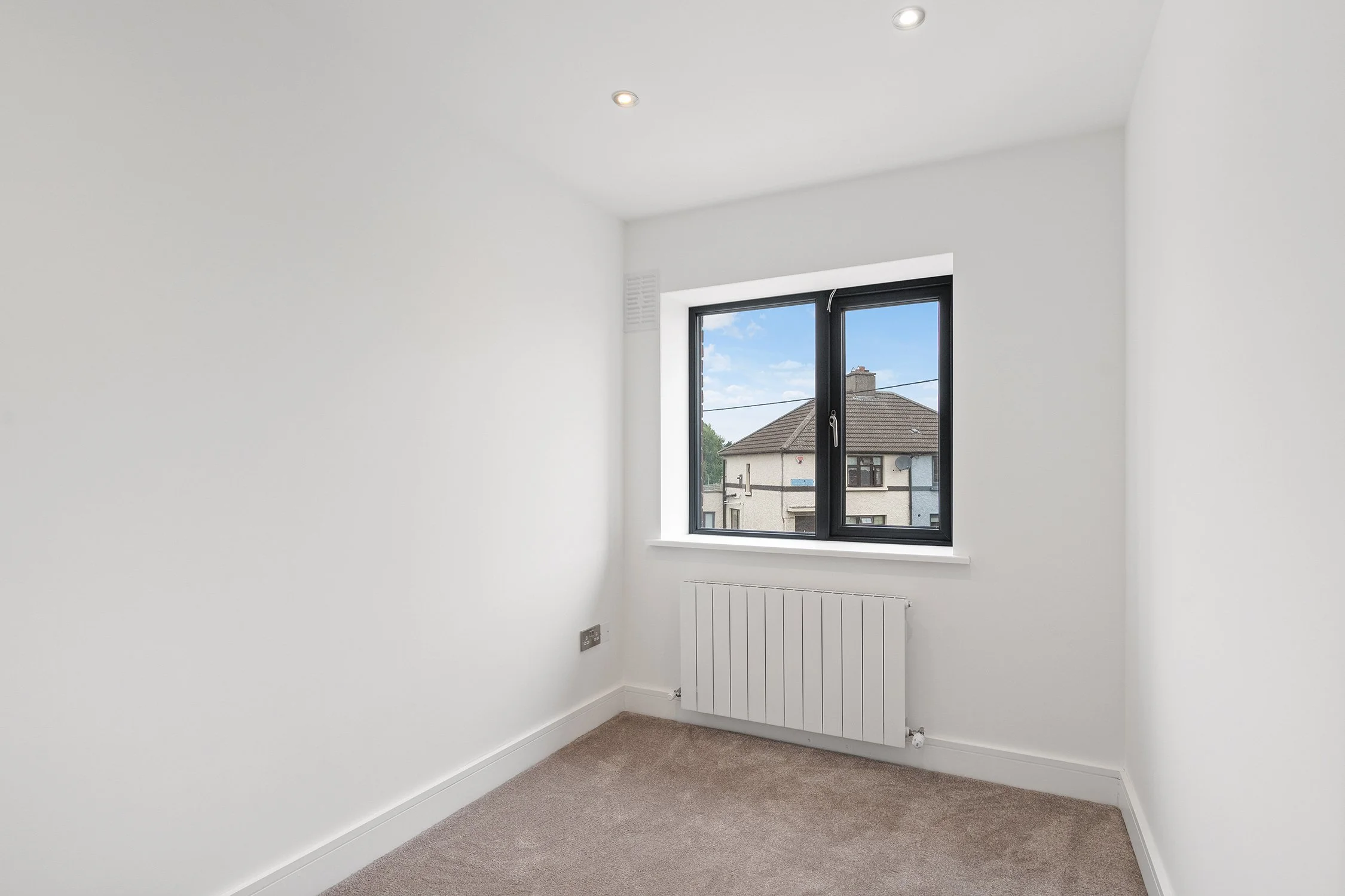 Empty white room with single window, beige carpet, white walls, and ceiling with recessed lighting.