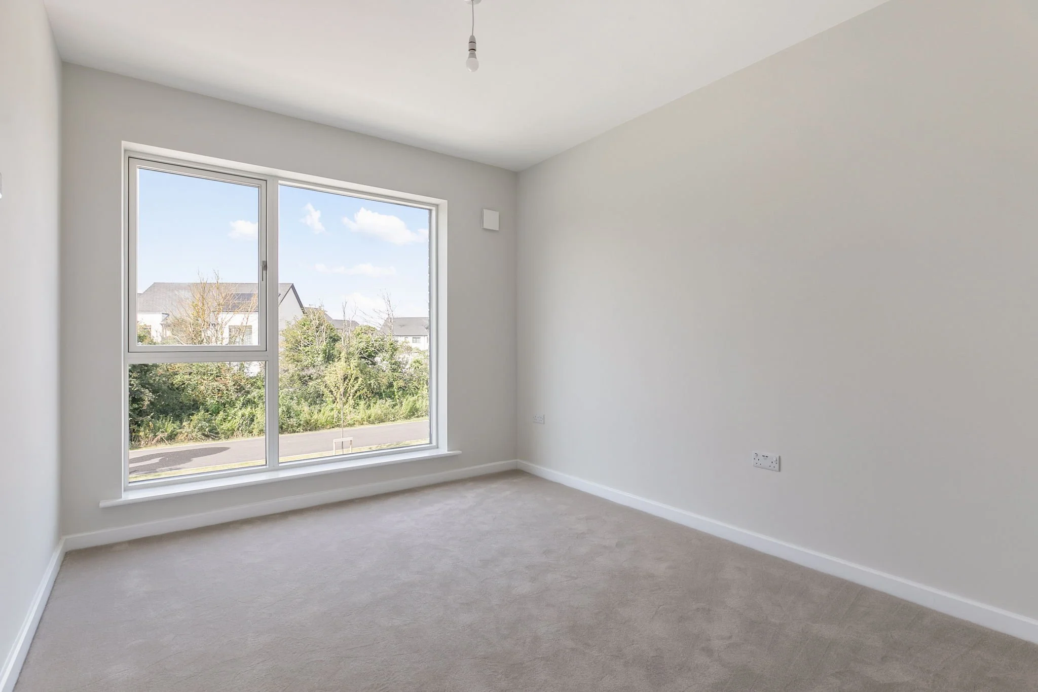 Empty room with a large window, white walls, carpeted floor, electrical outlets, and a ceiling light socket.