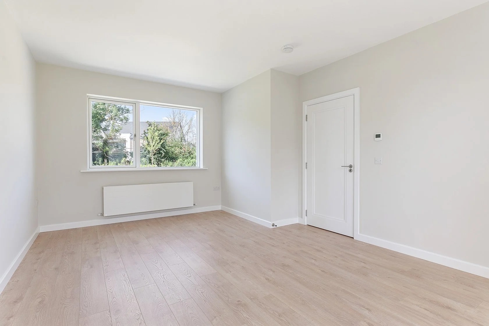 Empty room with light wood flooring, white walls, a window with a view of trees, a white radiator below the window, a white door, and a thermostat on the wall.