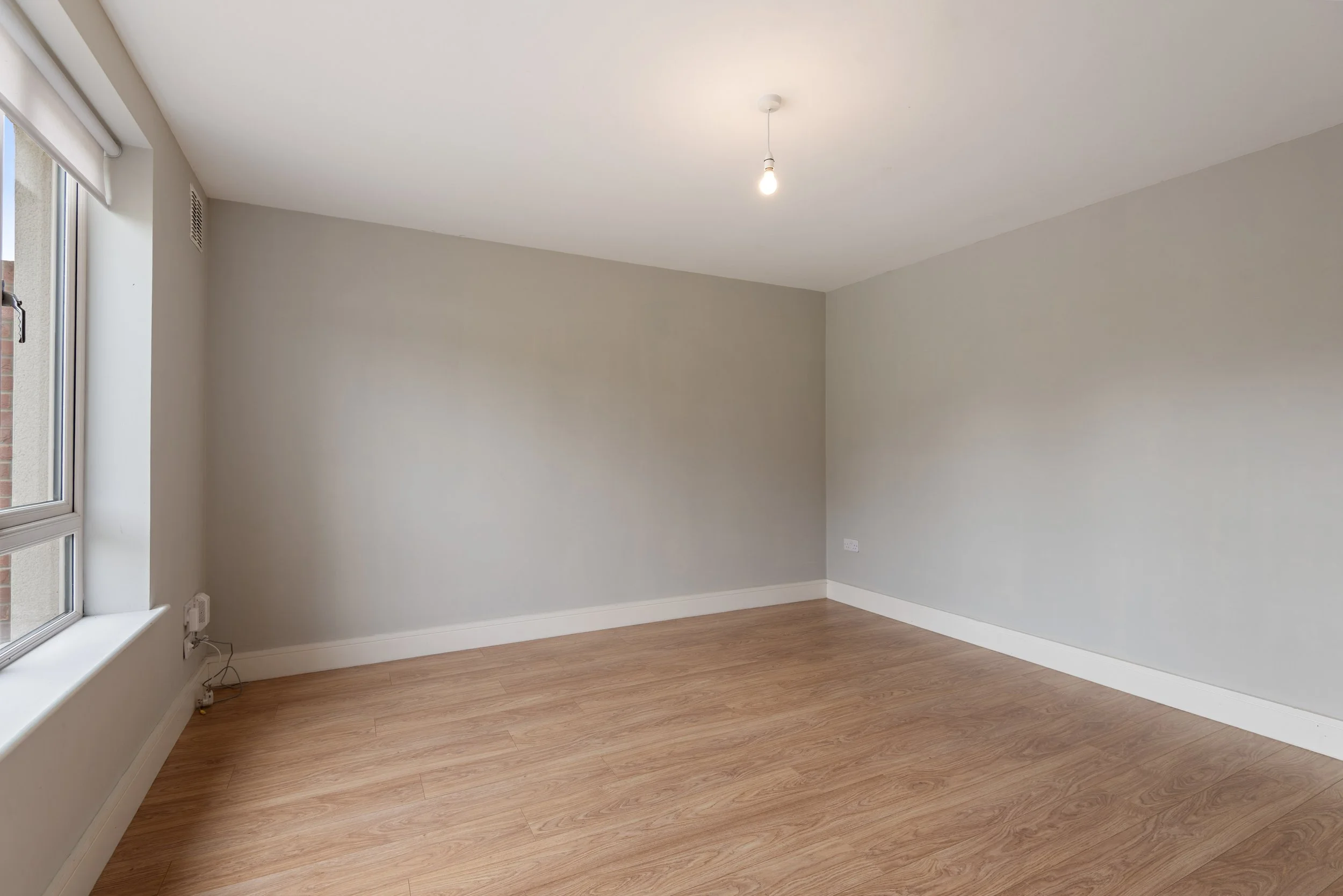 Empty room with light gray walls, a window on the left, a single light bulb hanging from the ceiling, and wooden flooring.
