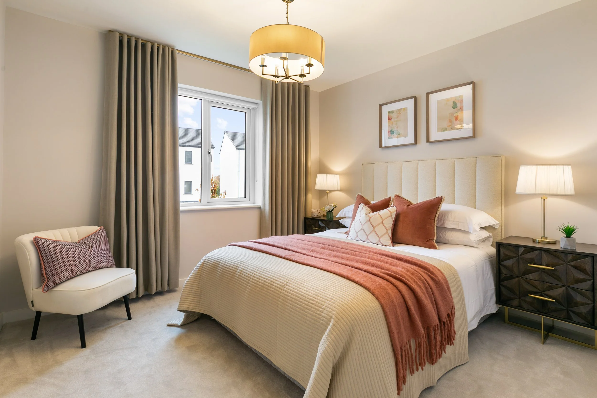 A bedroom with a large bed, beige upholstered headboard, and a cream-colored bedspread with rust-colored pillows and a throw blanket. There are two black nightstands with lamps, a white chair with a pillow, a window with beige curtains, and two framed art pieces above the bed.