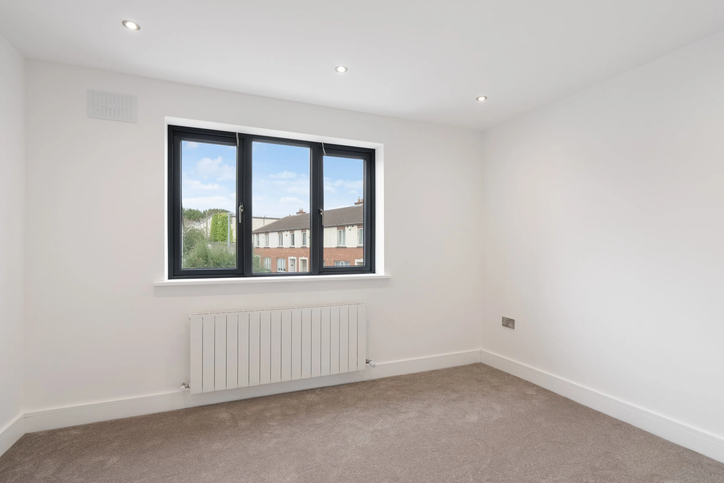 Empty room with white walls, beige carpet, black-framed window showing houses outside, and white ceiling with recessed lighting.