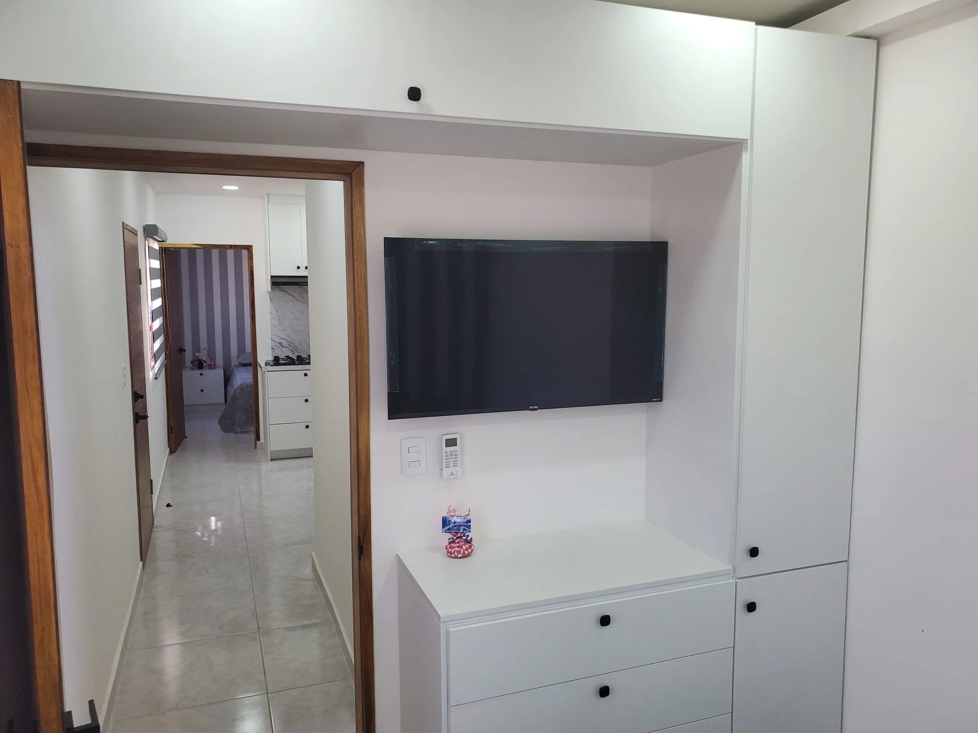 Living room with a wall-mounted flat-screen TV, a white storage cabinet with black knobs, and a hallway leading to bedrooms with tiled floors and striped curtains.