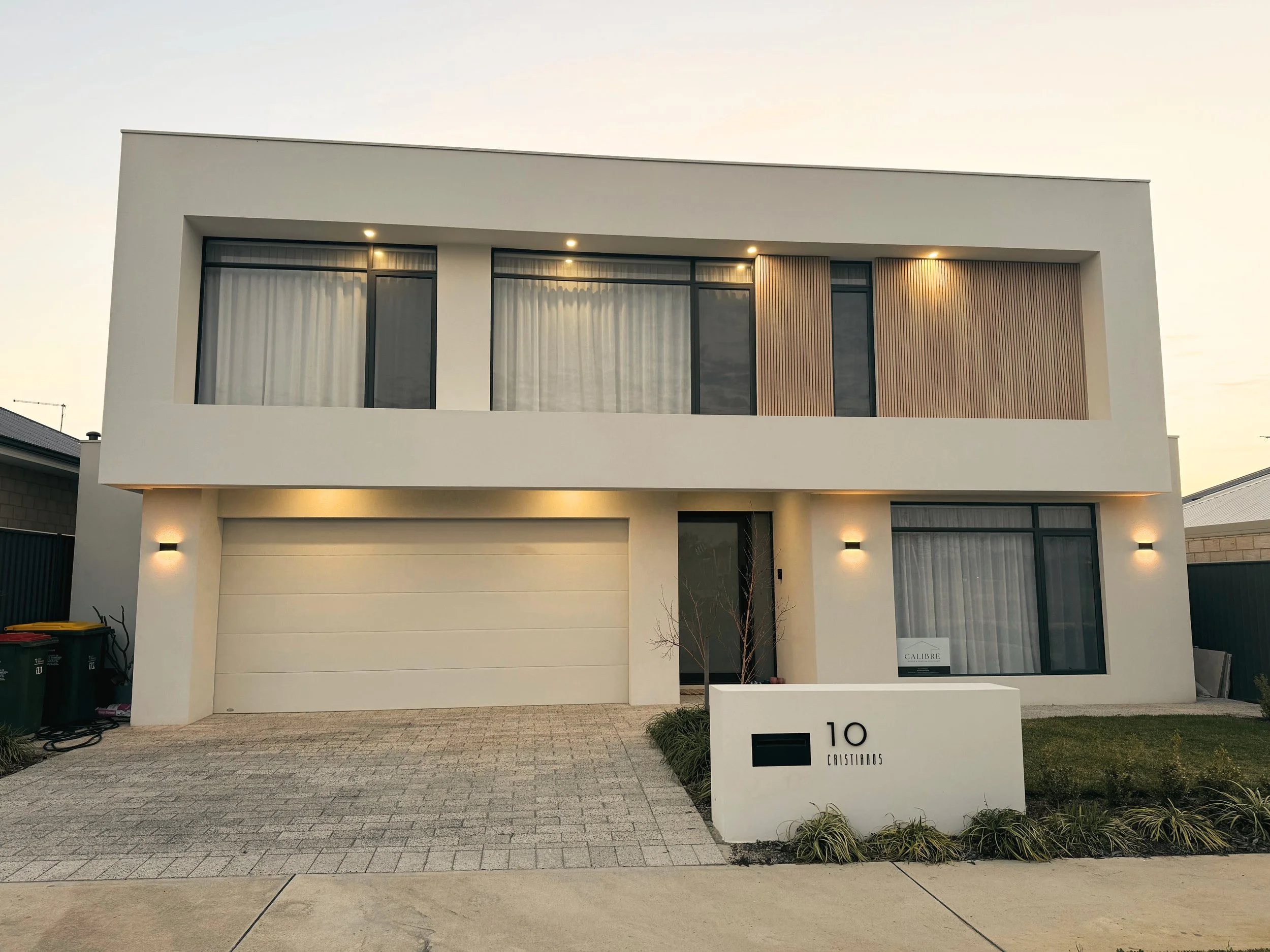 A modern two-story house with white exterior walls, large glass windows with curtain blinds, a wooden front door, and vertical wooden slats on a balcony. The house is surrounded by palm trees and desert plants, with a concrete driveway and landscaped lawn at sunset.