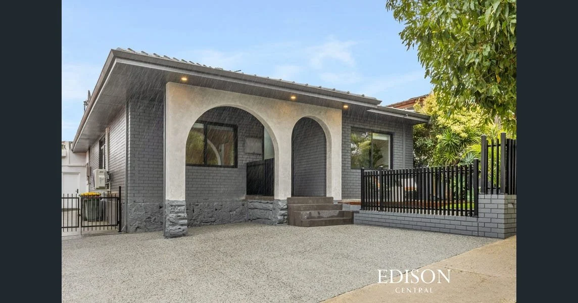 Front view of a modern house with a gray exterior, arched porch, large windows, steps, and a small fenced yard, under a blue sky with trees.