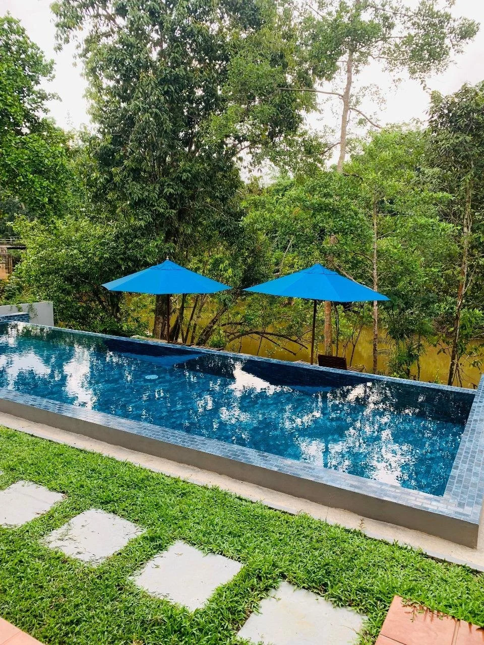 A swimming pool with two blue umbrellas beside a lush green area with tall trees and a small body of water in the background.
