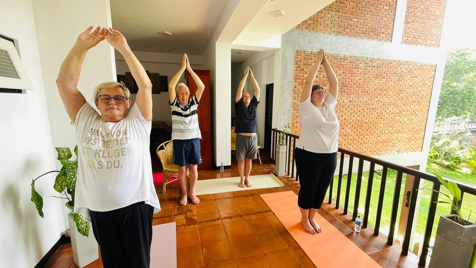 Four people practicing yoga on yoga mats on a balcony, with hands raised overhead in a prayer position.