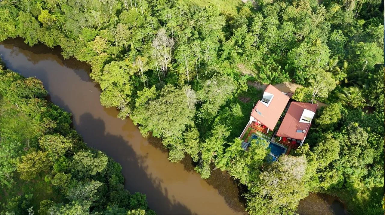 An aerial view of a house with a swimming pool surrounded by dense green trees near a muddy river.