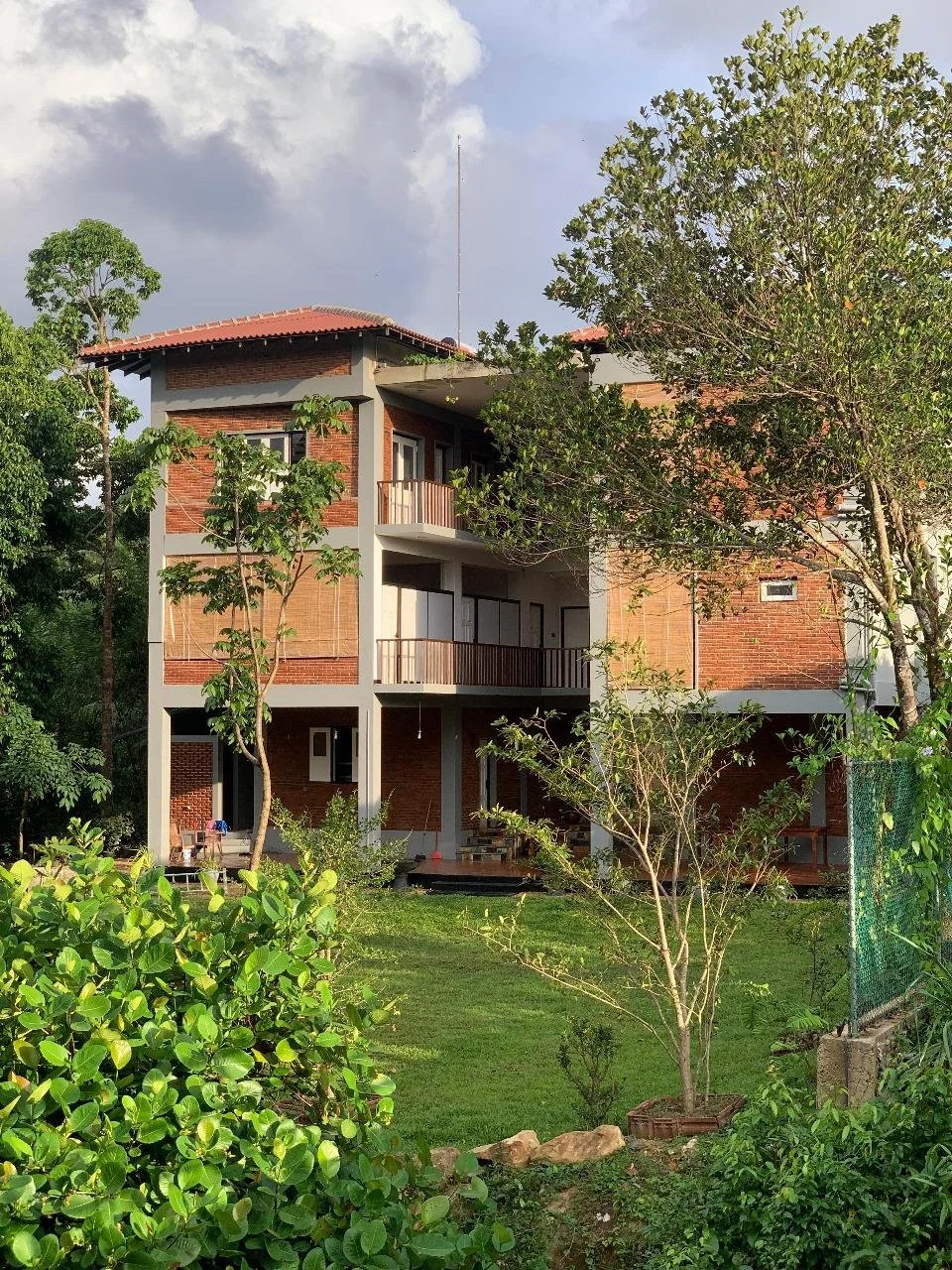 A multi-story house with brick walls, gray concrete accents, and a red-tiled roof seen through green trees and a garden.