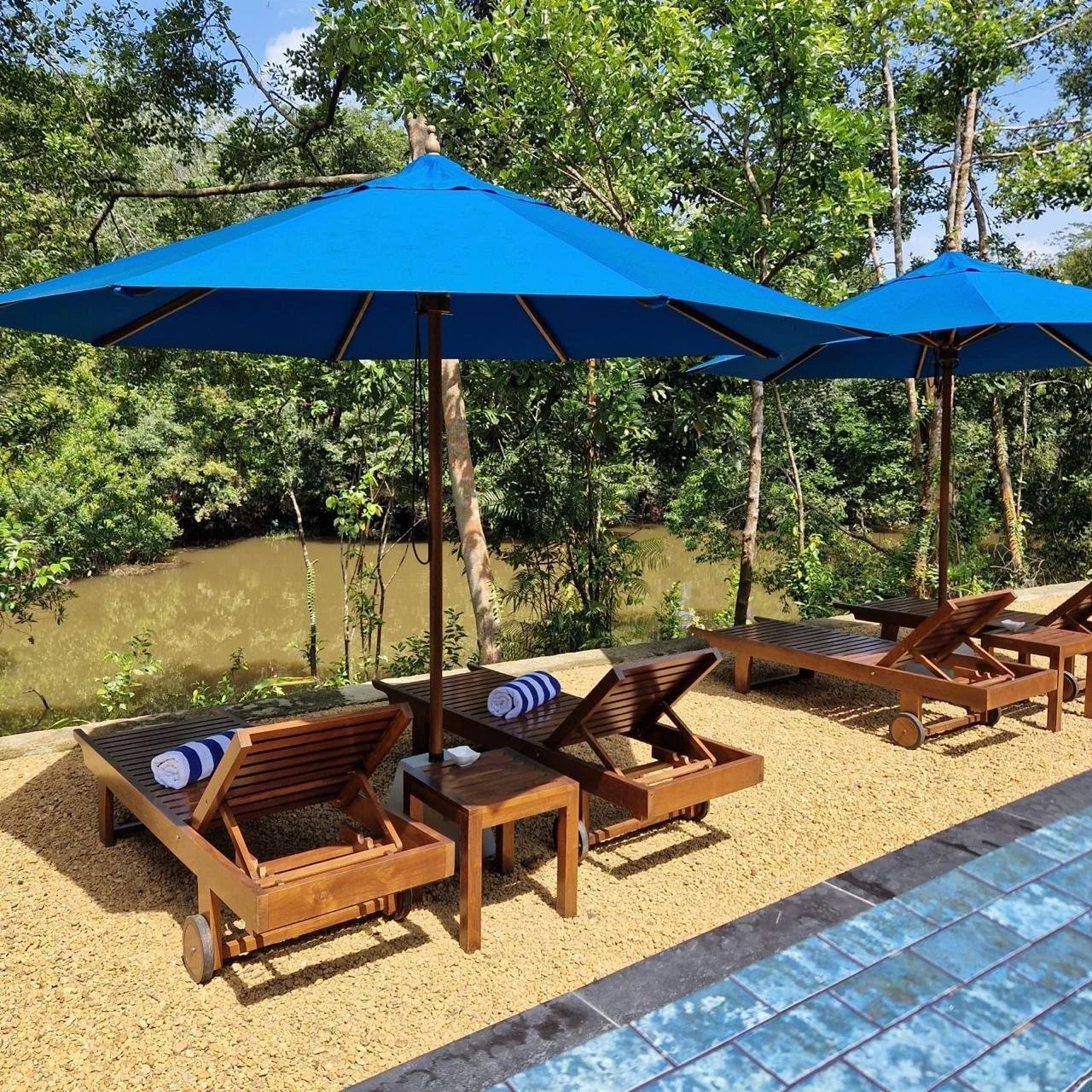 Three wooden lounge chairs with rolled towels on a yellow gravel poolside, shaded by blue umbrellas, overlooking a river and lush green trees.