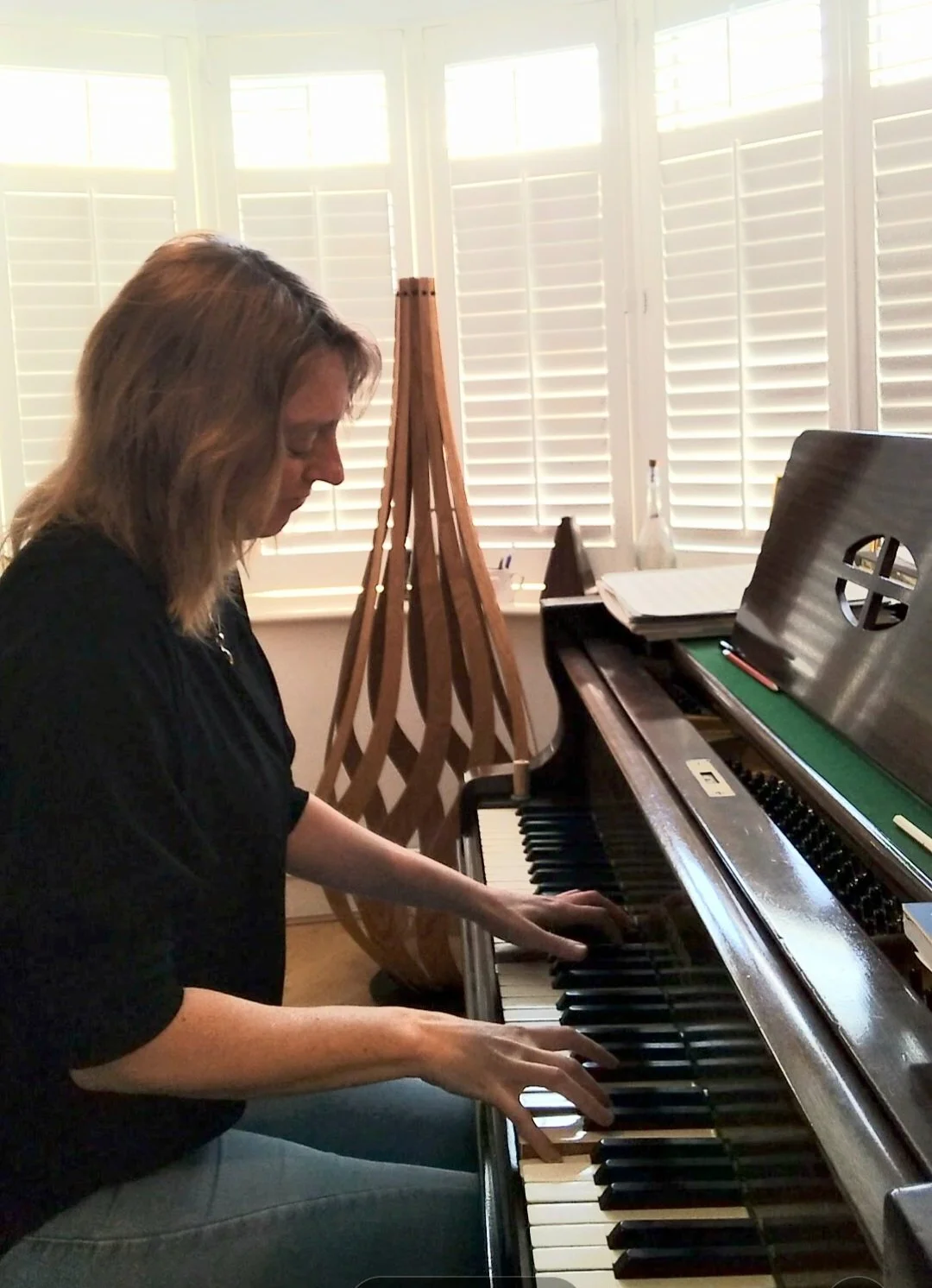 A woman playing the piano in a bright room with large windows and white blinds.