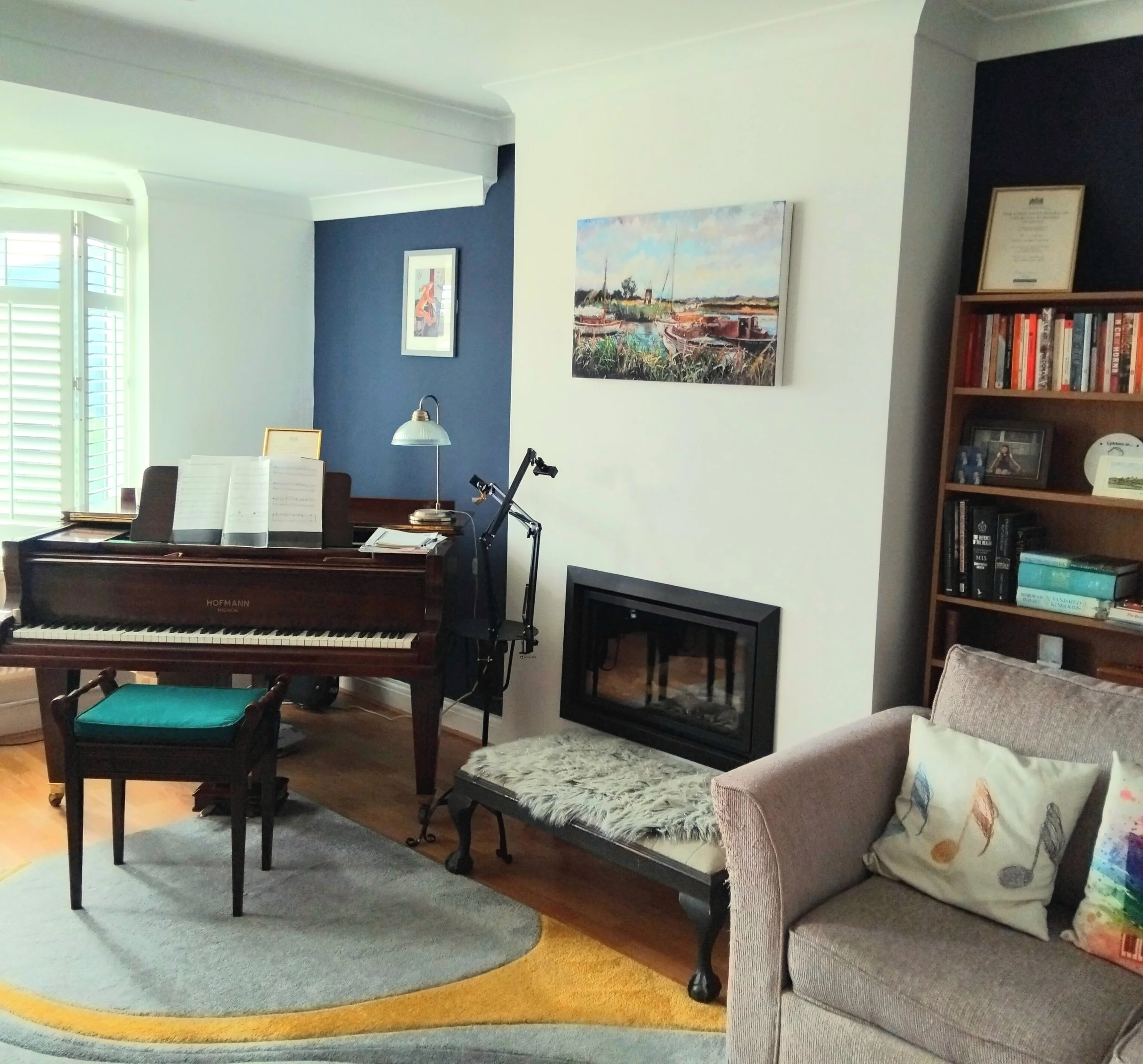 Living room with a brown upright piano, a matching piano bench, a gray armchair with colorful pillows, a carpet with a yellow border, a bookshelf, artwork on the wall, and a fireplace.