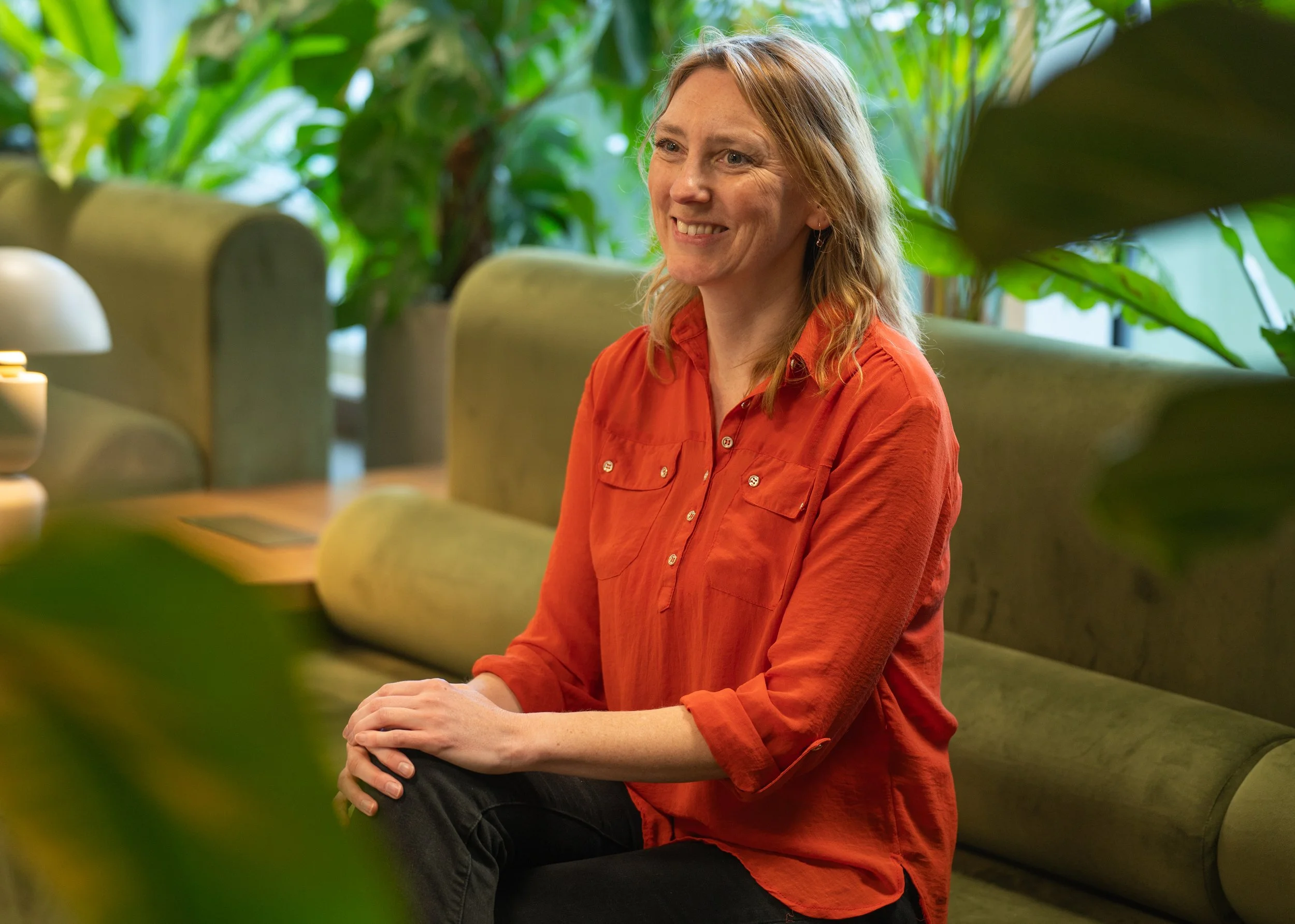 A woman sitting on a green sofa in a room filled with plants, smiling and wearing an orange shirt.
