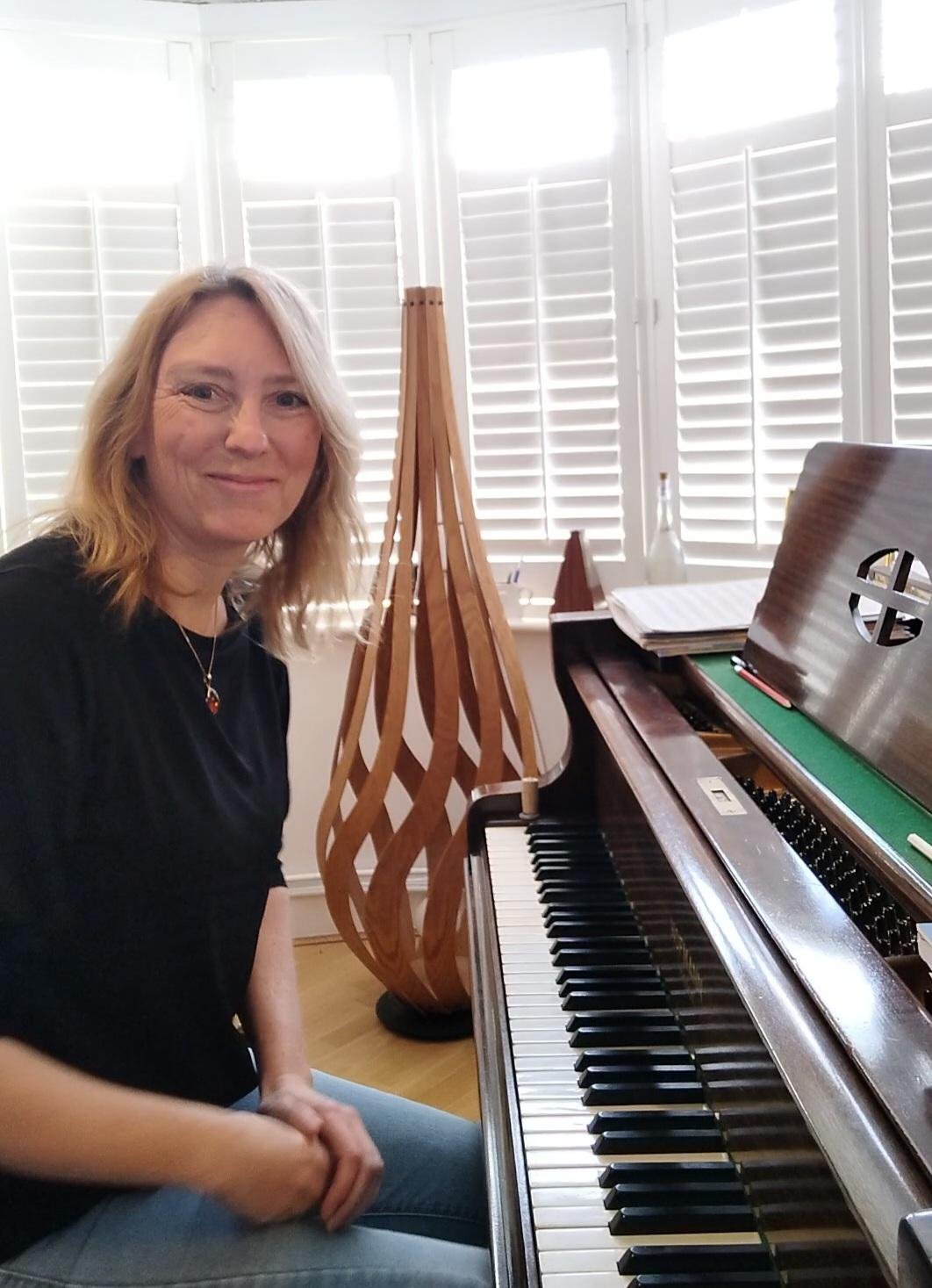 A woman sitting at a piano in a well-lit room with white window shutters, a wooden decorative lamp, and a music stand with sheet music.