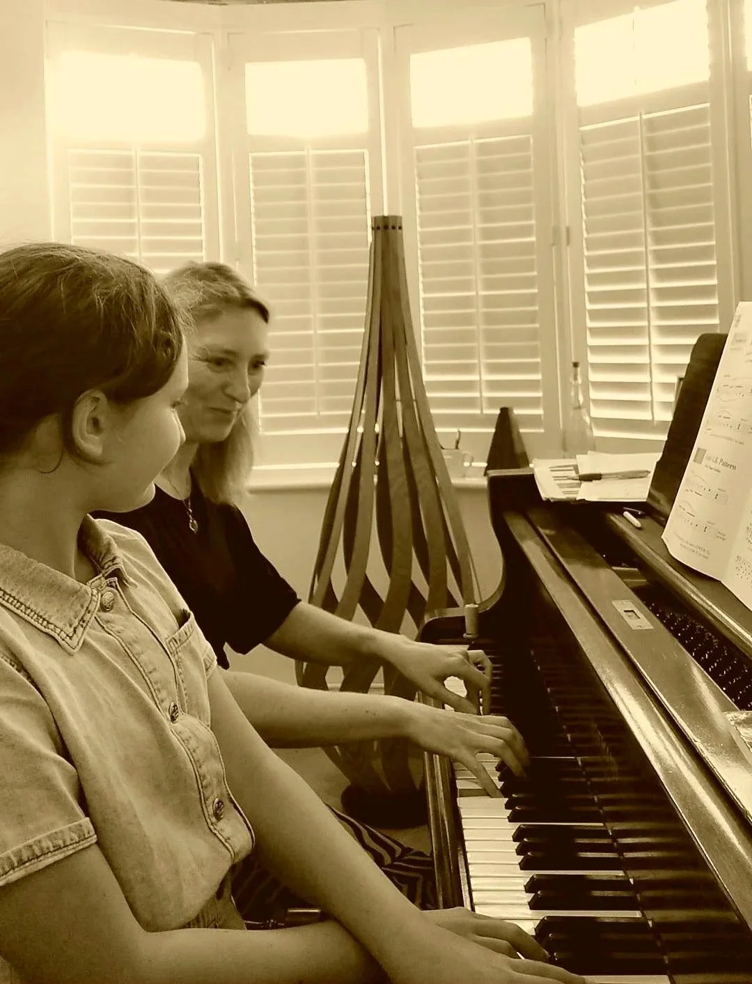 A piano teacher and student playing a piano duet together, reading sheet music, in a room with white shutters on windows and a wooden easel in the background.