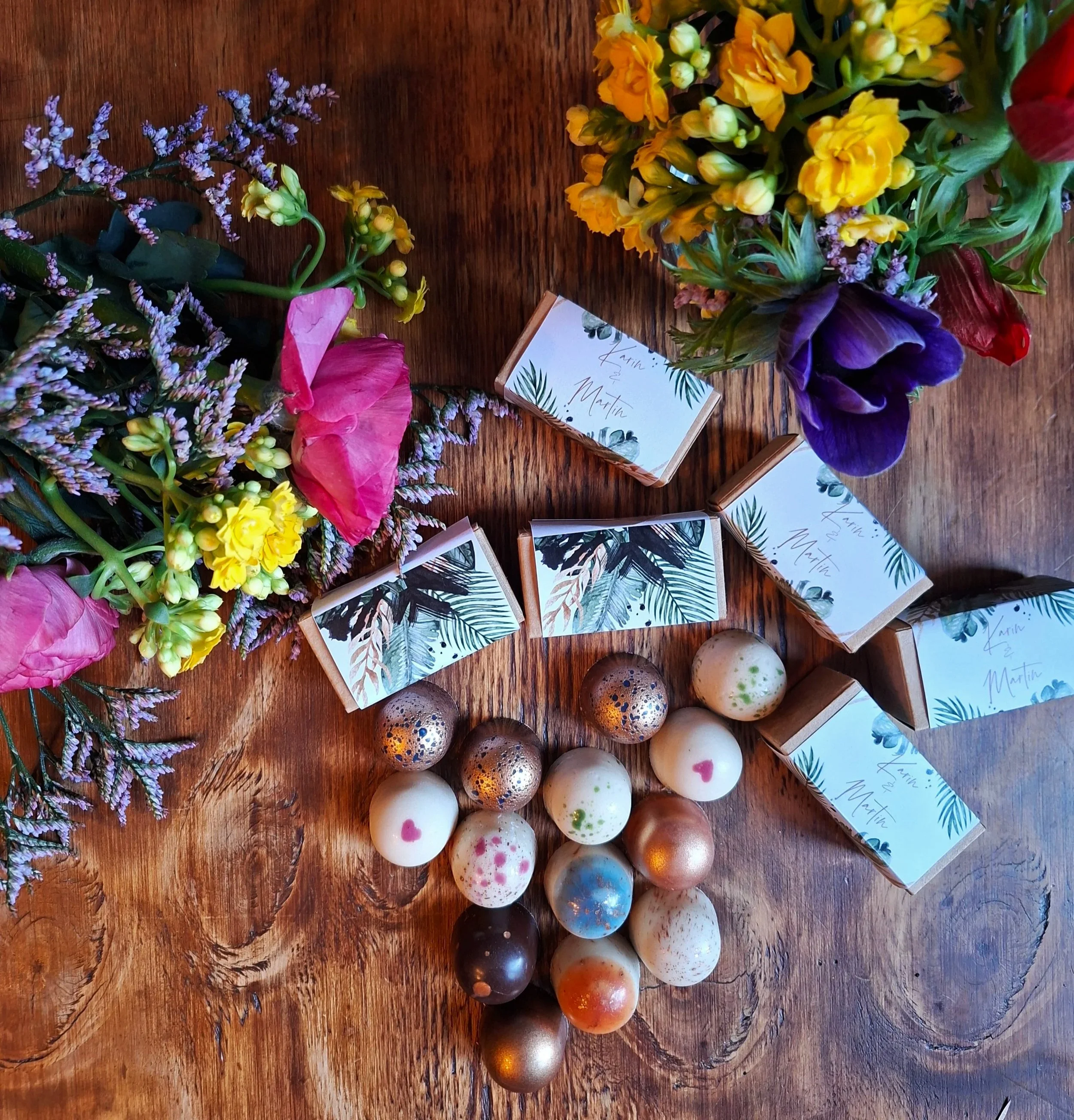 Aufsicht bunte Blumen mit bunten veganen Pralinen und kleinen Pappboxen mit bedruckter Banderolen. Zur Hochzeit