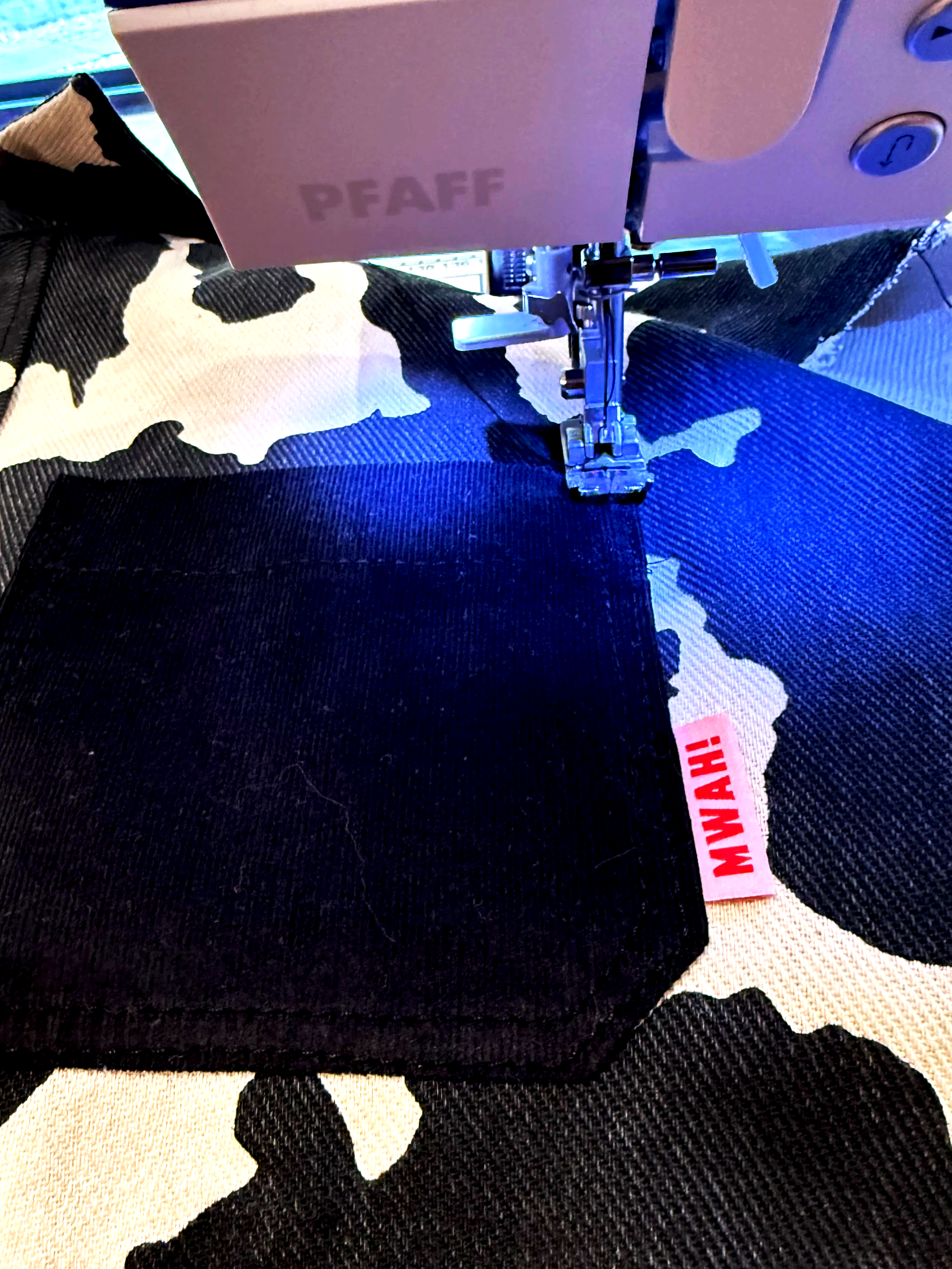 A sewing machine embroidering a black patch onto black and white camouflage fabric. The sewing machine has the brand name 'PFAFF' visible and a red label with 'WAH!' on the fabric.